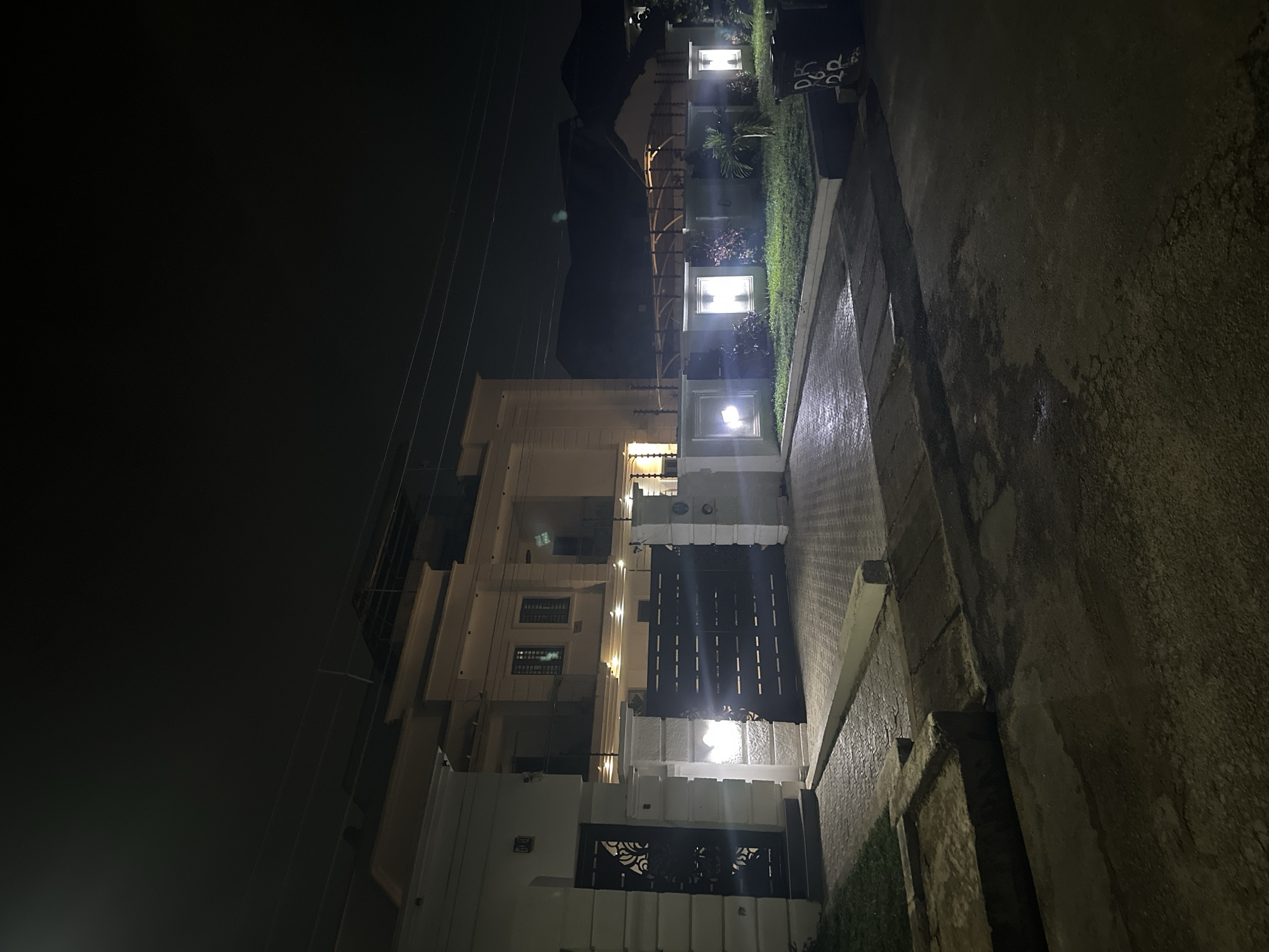 The image shows a night-time view of the exterior of a modern, multi-story residence in Abuja, Nigeria. The house is illuminated by artificial lights, highlighting its contemporary architectural design, which features clean lines, large windows, and a flat roof. A prominent dark metal gate, likely automated, is a central feature, leading to a driveway paved with interlocking bricks. Exterior lighting is strategically placed along the property's perimeter and in the garden area, which appears well-maintained with green grass and some vegetation. Overhead power lines are visible against the dark night sky. The overall scene suggests a secure and well-kept private property.