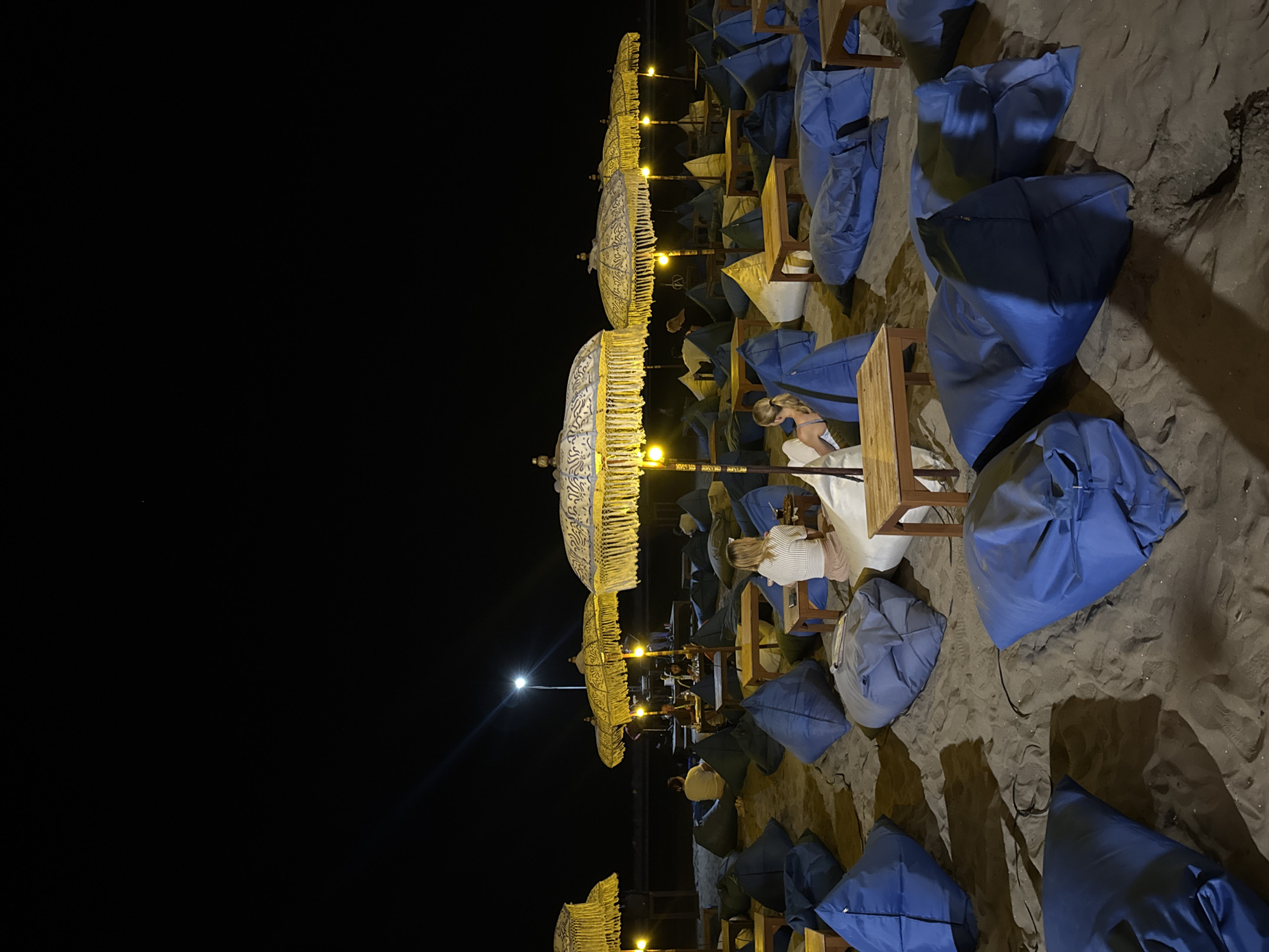 This is a night time photograph of a beachside seating area in Kuta, Indonesia. The foreground is filled with numerous large, blue beanbag chairs scattered across the sandy beach. In the mid-ground, several tables are situated between the beanbags, some with people seated at them. Above and behind these tables, a row of decorative, yellow-fringed umbrellas with intricate patterns are illuminated by soft lighting. Two women are visible, one seated at a table to the right of the frame, and another, partially obscured, seated further back. In the background, the dark expanse of the ocean meets the night sky, with a single bright light post visible near the horizon. The overall scene depicts a relaxed, outdoor dining or lounge setting.