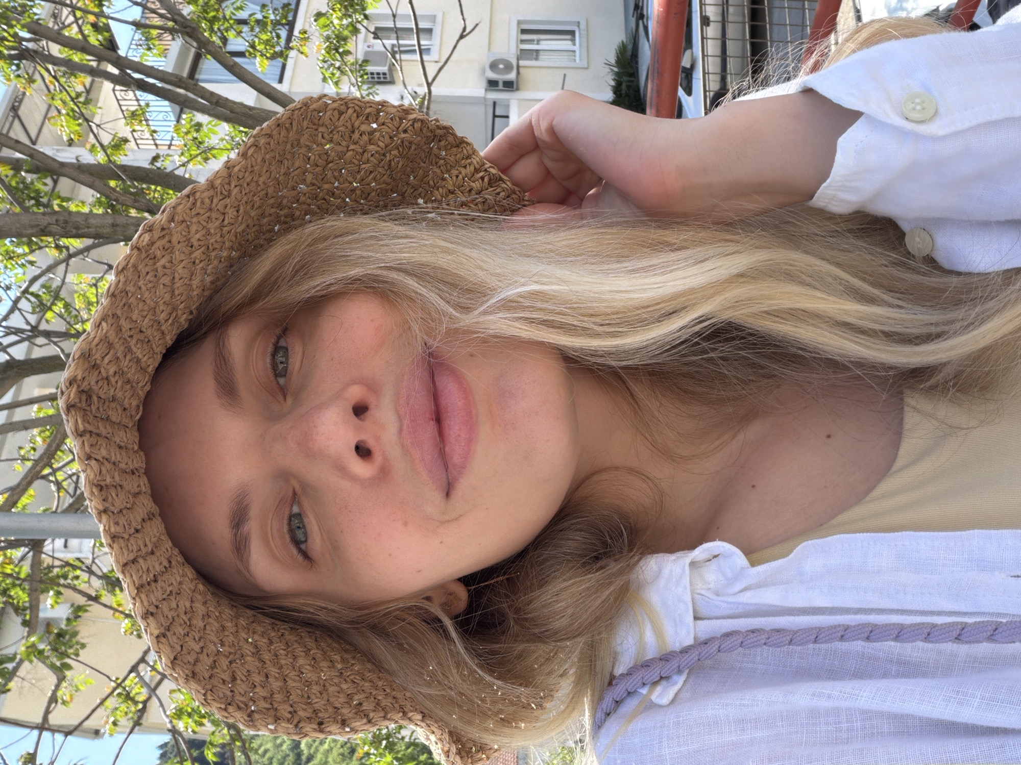 The image is a close-up, upward-facing selfie of a person outdoors in Budva, Montenegro, taken under bright daylight. The subject, appearing to be female, has long, wavy blonde hair and a fair complexion with visible freckles on her nose and cheeks. She wears a light brown straw hat, a white linen button-up shirt, and a light yellow top underneath. Her left hand is subtly raised, with fingers resting near the rim of her hat.
Her face is angled slightly towards the camera, with her green eyes looking directly upwards. Her lips are naturally pink and slightly parted. The background reveals several light-colored buildings with multiple windows, balconies, and visible air conditioning units, indicating an urban or residential environment. Deciduous trees with green leaves and bare branches are scattered in the background, suggesting a sunny, clear day. Metal railings from balconies or fences are also discernible. A braided strap, light purple or gray in color, possibly from a bag, hangs near her right shoulder. The overall impression is a casual, sunlit portrait. There is no visible text in the image.