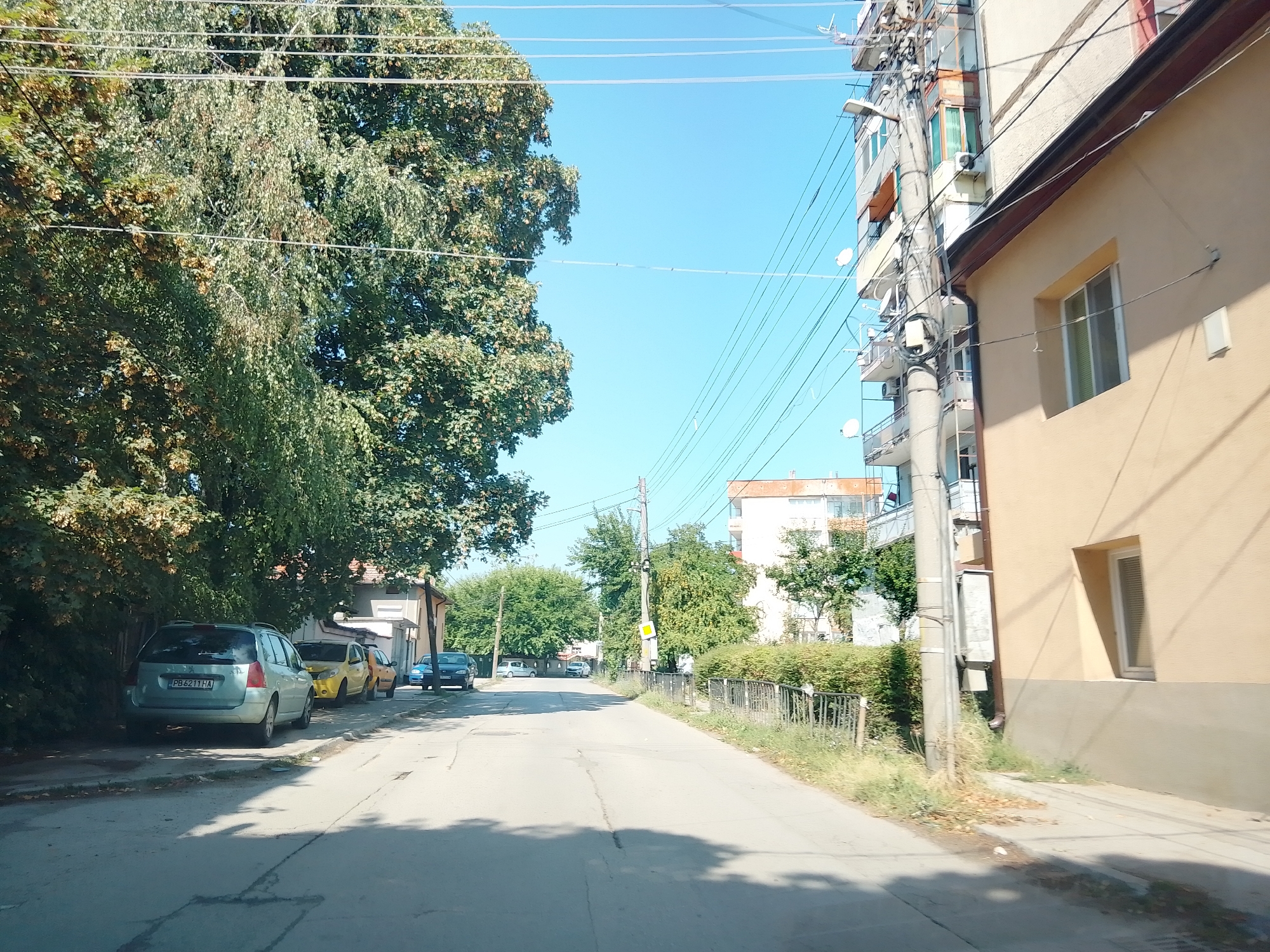 This is a street-level, eye-level, medium-range, outdoor, daylight, eye-level, full-shot photograph taken from a vehicle, looking down a paved street lined with buildings and trees. On the left side of the street, parked cars include a light blue station wagon with license plate "PB 6211 HA", a yellow car, and an orange car. Further down, a dark blue car is visible. A dense, leafy green tree occupies the left foreground, partially obscuring a light-colored building. On the right side of the street, a tan building with a single window and a drainpipe is prominent, with a tall utility pole and numerous overhead wires to its left. Behind the tan building, a multi-story residential building with balconies is visible. The street is a two-lane road with worn pavement and a shadow cast across it from the left. The sky is clear and blue. The location is identified as Ihtiman, Bulgaria.
