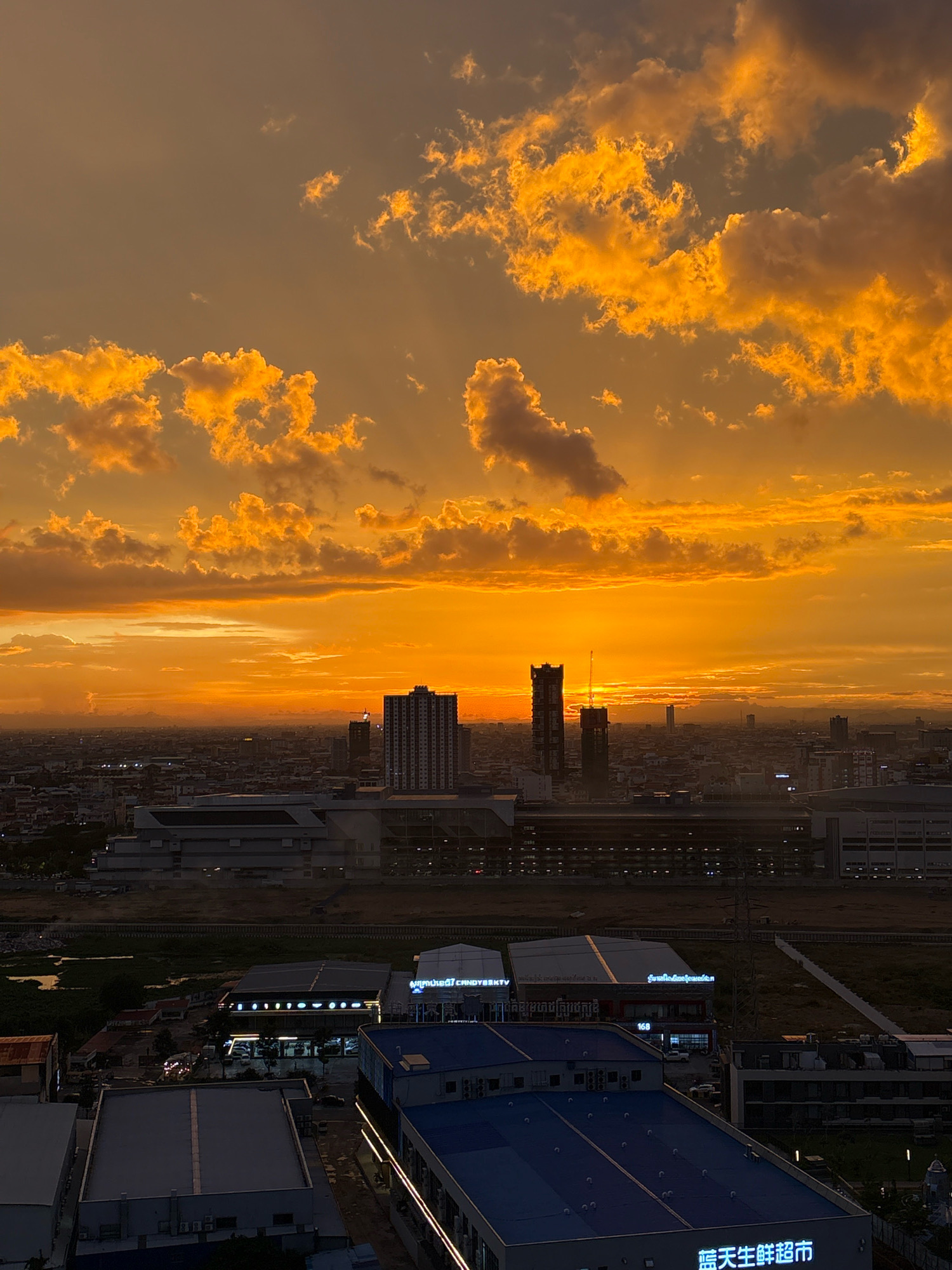 The photograph captures a sunset over a cityscape, likely Khan Mean Chey, Cambodia, characterized by a dramatic sky filled with fiery orange and yellow clouds. The sun is low on the horizon, casting a warm, intense glow that illuminates the clouds and the upper portions of the buildings.
In the foreground, several buildings are visible. To the left, a large, multi-story building with a flat, dark roof dominates the lower part of the frame. To its right, a smaller building with a blue roof and vertical white lighting accents runs along its facade. Further back, a building with a darker roof displays illuminated signage, including the characters "168" and what appears to be text in Khmer. Next to it, another building has illuminated signage that reads "AMUSEMENT CANDY". In the middle ground, a large, modern structure with a light-colored facade and a dark, sloped roof stretches across the image.
The middle distance features a dense urban landscape with numerous buildings of varying heights. Prominent among these are two tall, dark skyscrapers; one is significantly taller and appears to have a construction crane on top. To the right of these skyscrapers, a lighter, white-and-grey high-rise building stands out, its windows reflecting the sunset light. The cityscape extends to the horizon, where a hazy layer of mountains or distant buildings can be discerned against the bright sunset.
The sky is the most striking element of the image. It is a gradient of deep orange at the horizon, transitioning to bright yellow and then to a lighter, dusky orange higher up. Fluffy cumulus clouds are scattered throughout the sky, catching the sunset's light and appearing as vibrant, glowing masses of orange and yellow, with some darker grey shadows.
The overall impression is one of a city at dusk, bathed in the spectacular colors of a setting sun. The scene is devoid of visible people or specific actions, focusing instead on the architectural elements of the city and the dramatic natural phenomenon of the sunset. The lighting is low, suggesting it is late afternoon or early evening.