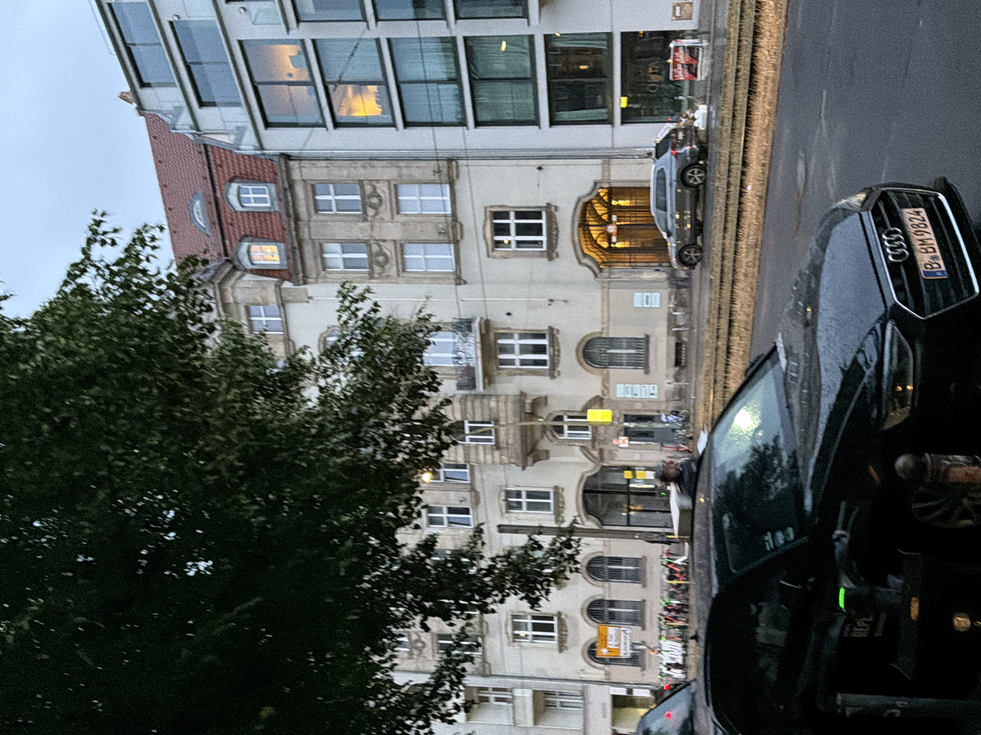 An overcast day in Berlin, Germany, with a tree in the foreground obscuring part of the view. In the middle ground, a row of buildings lines a street. The building on the left is older, with a mansard roof and ornate architectural details. Next to it is a modern building with large glass windows and balconies. The street appears to be a tram line, with tracks visible. Several vehicles are present: a black Audi in the foreground with its license plate visible (B.BM9824), and a gray SUV parked on the side of the street. Scooters are lined up along the sidewalk. The sky is cloudy, and the lighting suggests it is late afternoon or early evening.