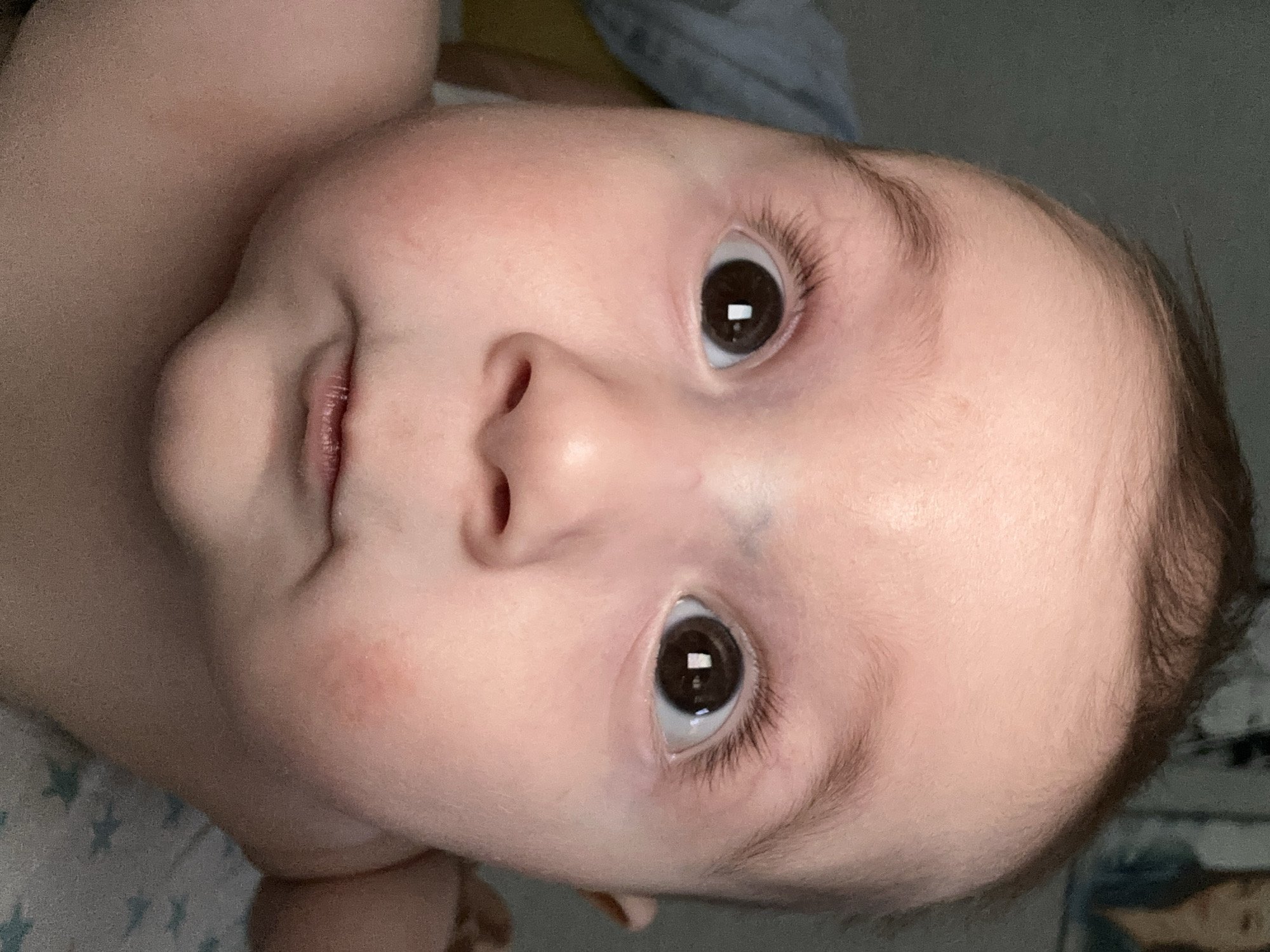 A close-up, slightly rotated, image features the face of an infant. The baby has fair skin, brown hair, and brown eyes. The background is blurred, showing a bed with patterned sheets. There is no indication of the location being Chaikovsky, Russia.