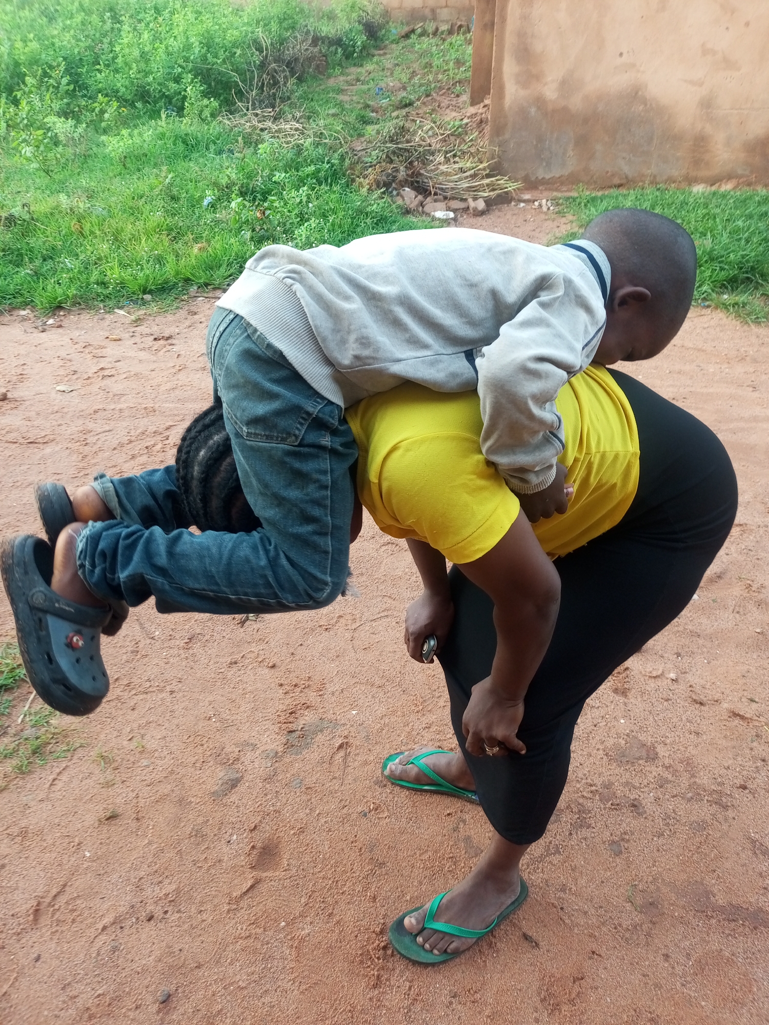 In the city of Tempure, Nigeria, a young child, dressed in a grey long-sleeved shirt and blue jeans, is being carried on the back of an adult. The adult is wearing a bright yellow t-shirt and black leggings, with green flip-flops on their feet. The child is positioned head-down, with their legs dangling. The scene takes place outdoors on a dirt path, with sparse green vegetation and a building visible in the background. The overall impression is one of playful interaction between the two individuals.