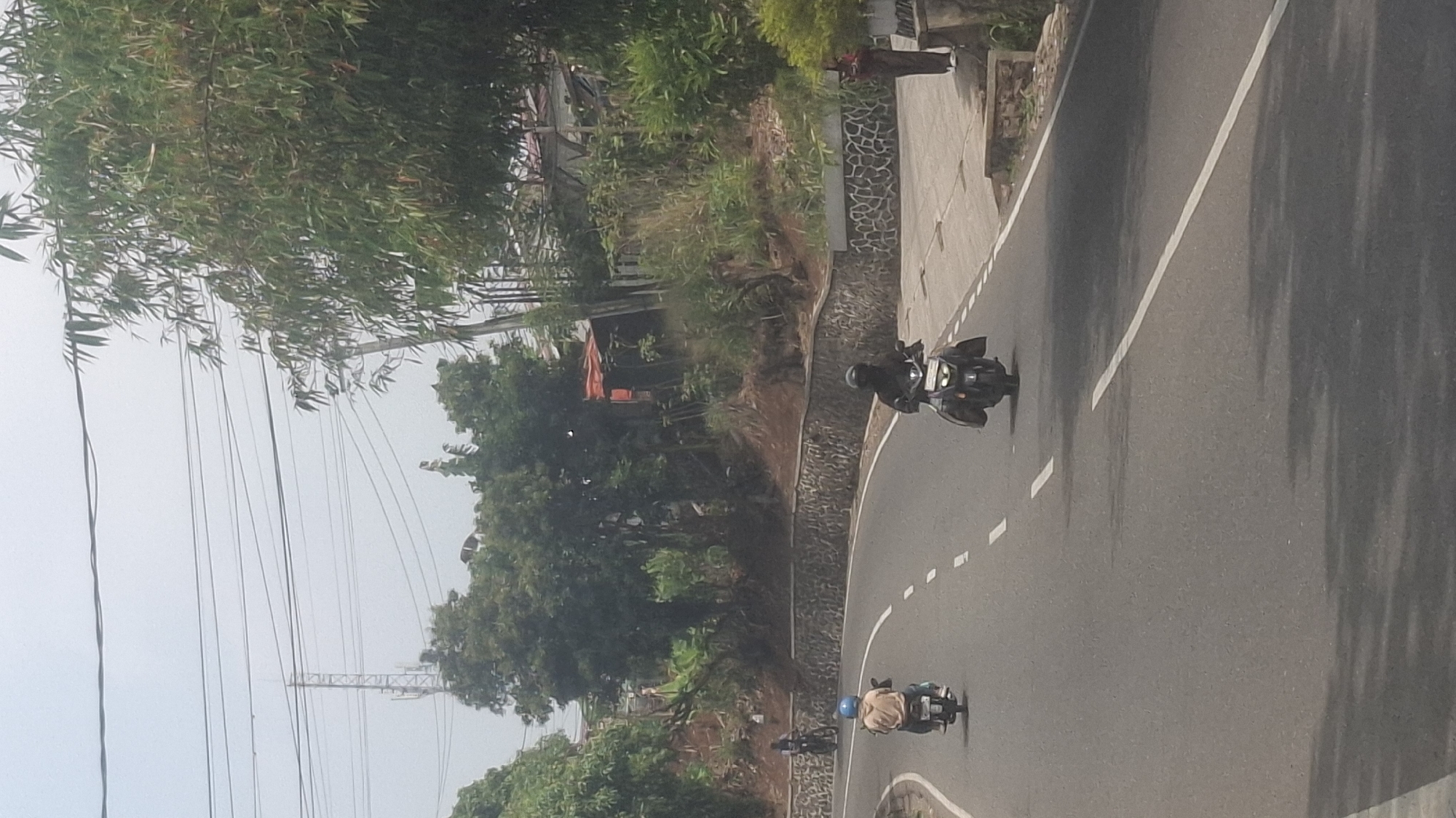 In Sirnagalih, Indonesia, a road curves gently through a landscape bordered by lush vegetation and a stone retaining wall. Two motorcycles are visible on the asphalt road, each carrying a rider. The motorcycle in the foreground has a rider wearing dark clothing and a helmet. Ahead of it, another motorcycle with a rider wearing a light-colored jacket and a blue helmet is also traveling on the road. The environment includes trees, a power line tower in the distance, and some buildings nestled among the foliage on the hillside. A person in a reddish-brown outfit is standing on the right side of the road, near a low wall. The sky is overcast.