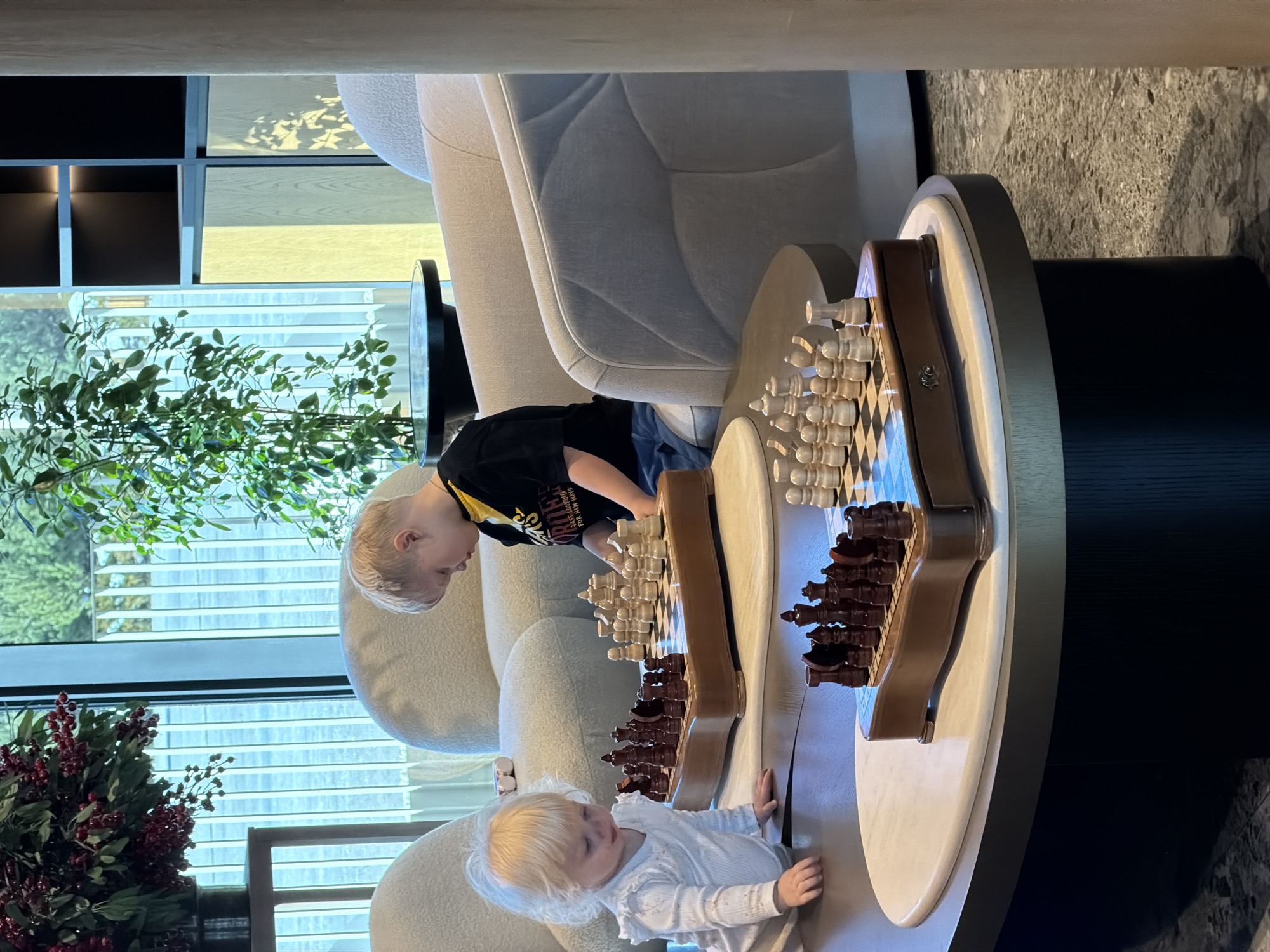Two young children are observing a chess board atop a round table. One child, wearing a black t-shirt, is leaning over the table and looking down at the chess pieces. The other child, wearing a white shirt, is reaching towards the chess board with their hands. The table is positioned in a room with a large window, a plant, and a sofa. The location of this scene cannot be confidently identified as being in Озёрное, Russia.