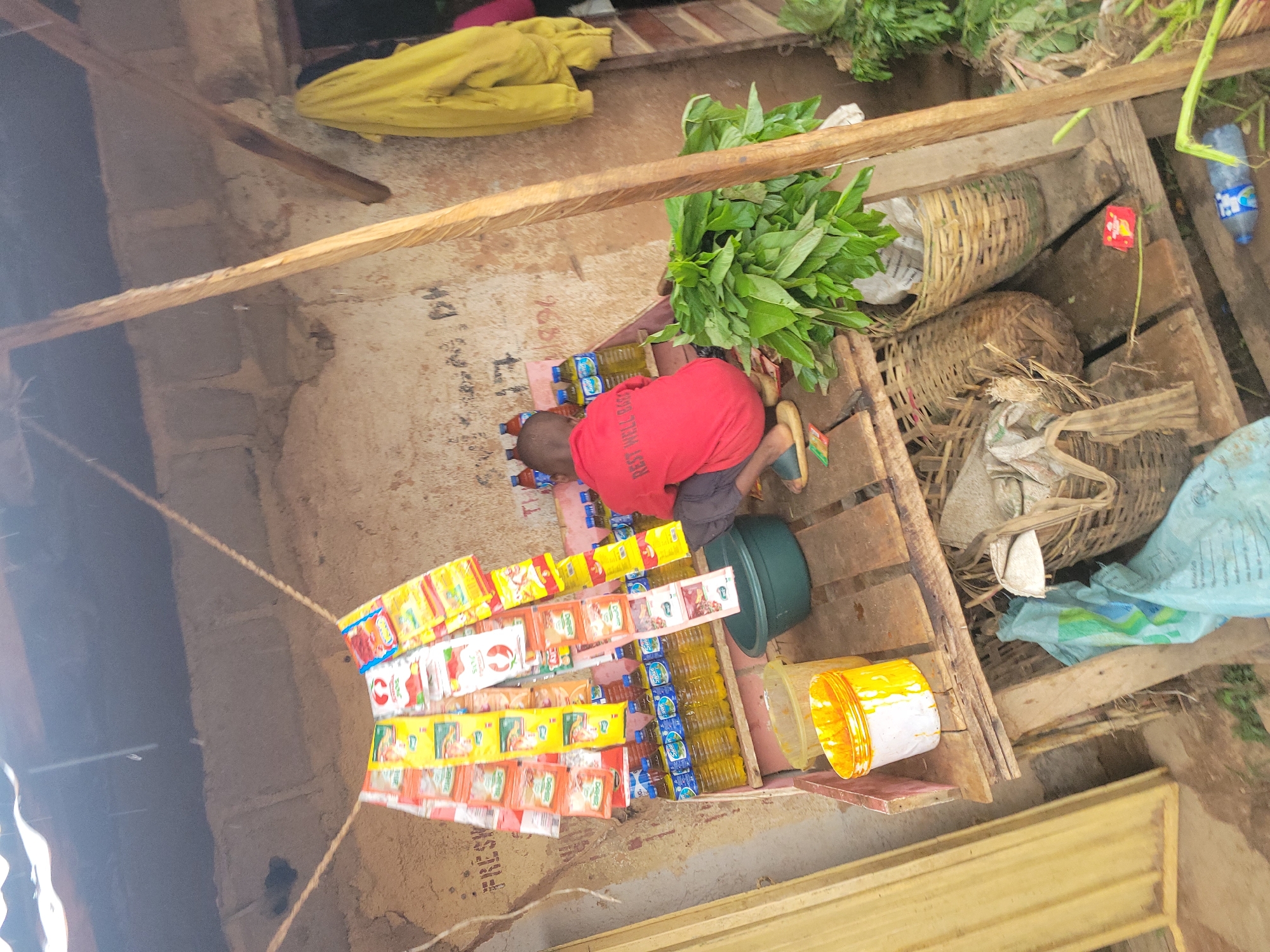 The image depicts a makeshift outdoor market stall in Jos, Nigeria. A young boy, wearing a red t-shirt with the text "REST WELL BABE" visible, is crouched down amidst the merchandise. The stall features a wooden shelf laden with bottles of yellow liquid, likely cooking oil or beverages, and a display of colorful packets of spices or seasoning hanging from a rope. To the left, a yellow bucket overflows with a viscous orange substance, possibly palm oil. A dark green bowl sits on a lower wooden platform. Behind the boy, a pile of fresh green leafy vegetables is bundled in a woven basket. A yellow jacket hangs from the wall of a nearby structure. The overall environment suggests a modest, informal trading post within a community setting.