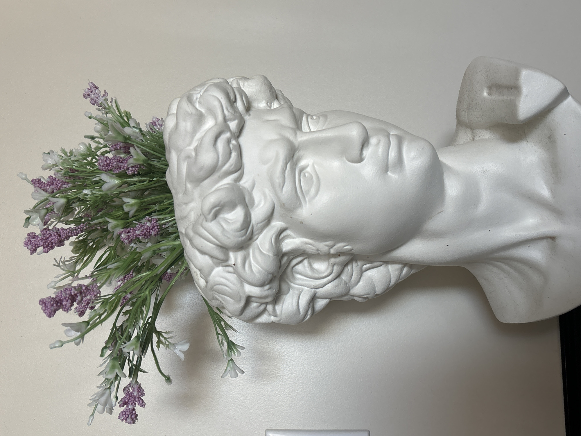 A white, plaster bust of a male head, likely a replica of a classical sculpture, lies on its side against a cream-colored wall. A bunch of artificial flowers, including purple lavender and white filler flowers, protrudes from the back of the head, suggesting it is being used as a vase or decoration. The bust is positioned close to the wall, with a portion of its base or support visible on the right side of the frame. The overall scene appears to be indoors. Location details, including the city and its known name, are unspecified.