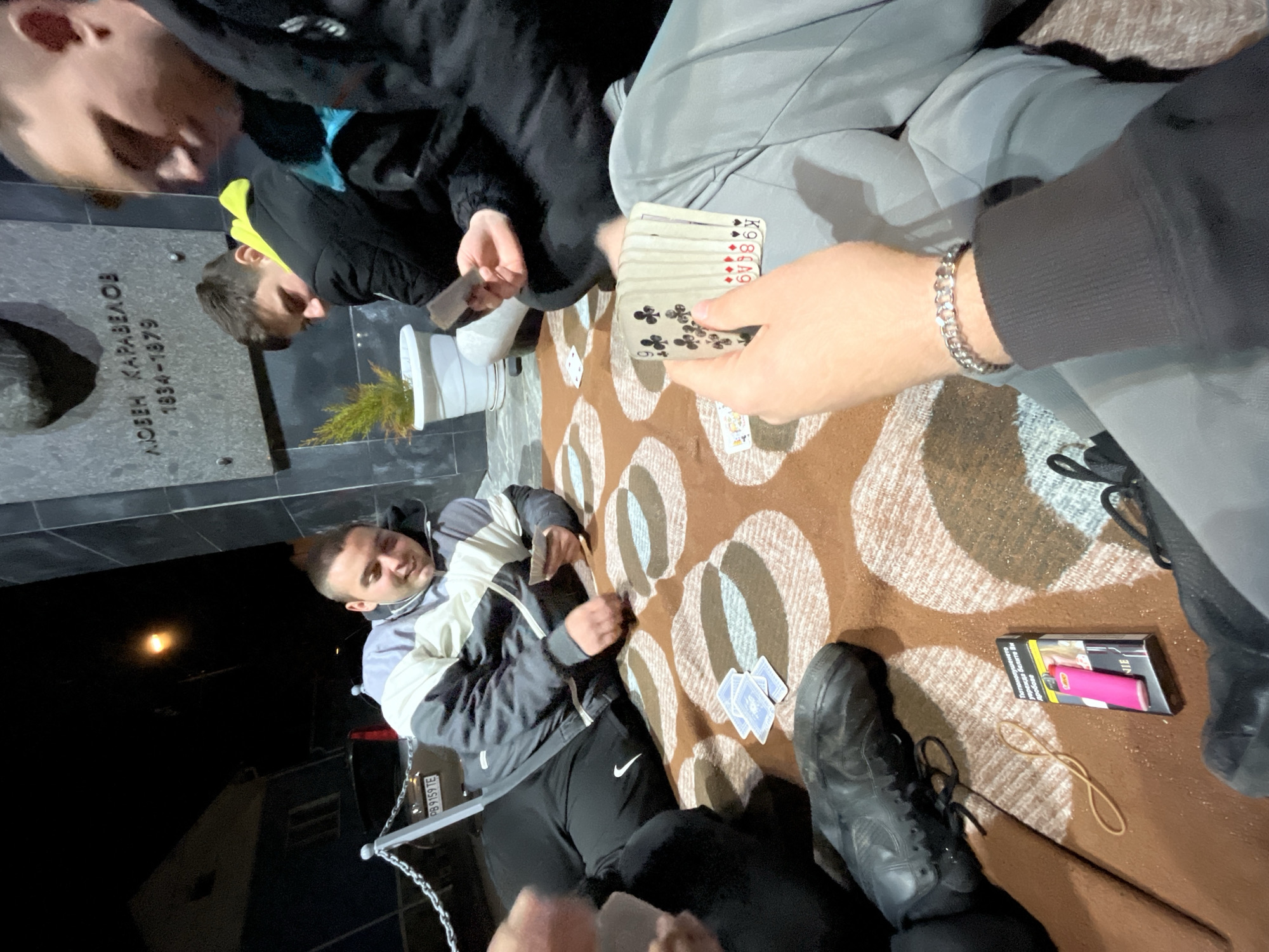 The image shows a group of people playing cards outdoors at night, seemingly near a monument or grave marker with Cyrillic inscriptions ("ЛЮБЕН КАРАВЕЛОВ 1834-1879"). This indicates a potential location in Karavelovo, Bulgaria, associated with Lyuben Karavelov, a Bulgarian writer and revolutionary. Four people are visible, though not completely, around a patterned blanket on the ground, which serves as their playing surface. Cards are in their hands and scattered on the blanket. One person holds a stack of cards, prominently displaying the clubs suit. A pack of cigarettes, a rubber band, and a pair of black shoes are also on the blanket. One man wears a grey and black jacket with black trousers and Nike logo on the pants. A car with a Bulgarian license plate ("PB 9159 TE") is partially visible in the background. The lighting is dim, with visible artificial light sources.