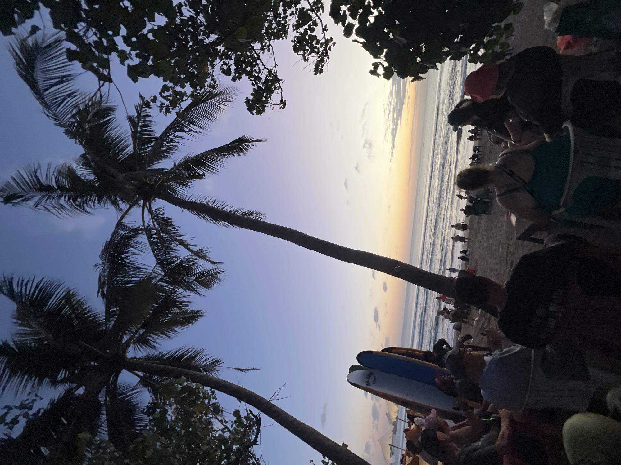 The image captures a tranquil sunset scene on Kuta Beach, Indonesia, where a group of people is gathered on the sand. Tall palm trees frame the view, their fronds silhouetted against the twilight sky. In the foreground, several surfboards are propped up, suggesting a surfing locale. The ocean stretches to the horizon, with gentle waves lapping the shore. People are seated in chairs, some facing the sea, others in conversation. The atmosphere is relaxed and peaceful, with the soft glow of the setting sun casting a warm hue over the beach. The beachgoers appear to be enjoying the end of the day, with the sound of the waves and the cool sea breeze providing a serene backdrop.