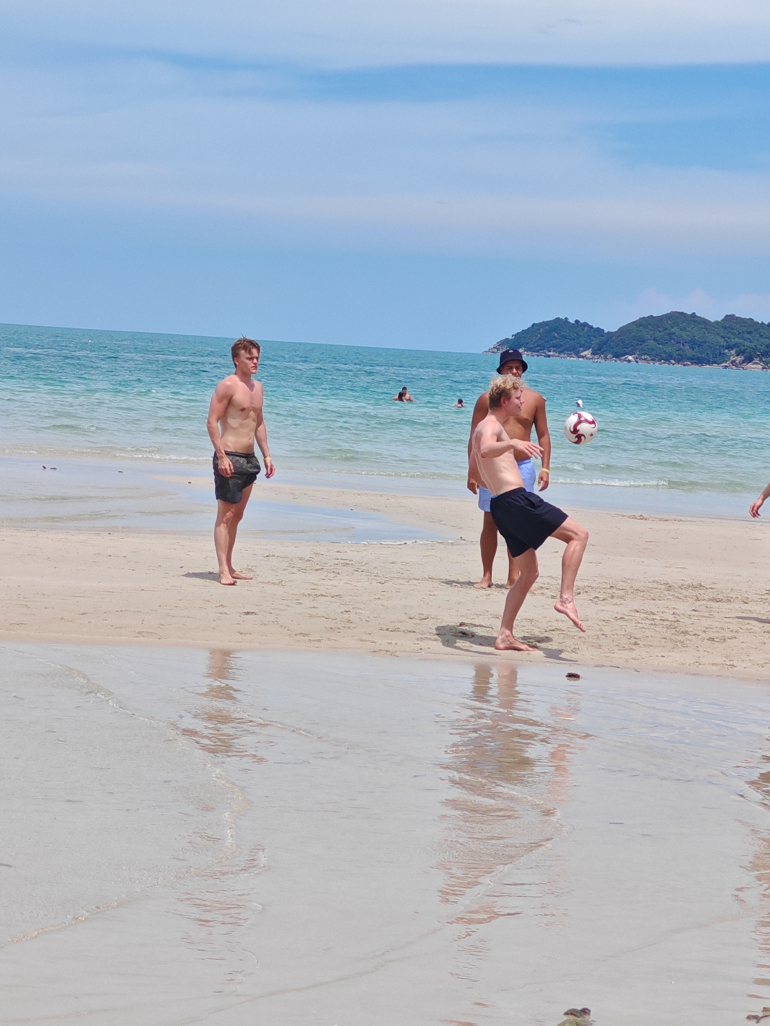 In Baan Chaweng, Thailand, on a sandy beach, three young men are engaged in a game of soccer. The man on the left, wearing dark shorts, stands on the sand. In the center, another man, also in dark shorts, is captured in mid-air, kicking a soccer ball with his right foot. Behind him, a third man, wearing blue shorts and a black hat, observes the play. The ocean with gentle waves stretches to the horizon, and distant green islands are visible under a bright, cloudy sky. The sand is wet and reflective near the water's edge, showing ripples and reflections of the sky and the people.
