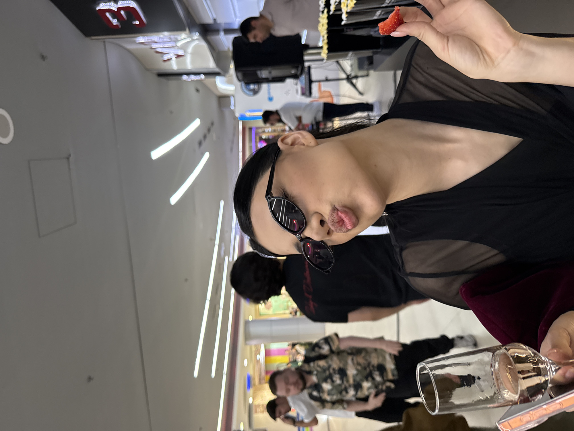 The image captures a scene indoors, likely in a shopping mall, with a person in the foreground holding a strawberry and a champagne glass. This individual is wearing dark sunglasses and a black outfit with sheer elements. The person appears to be posing, possibly taking a selfie. Several other people are visible in the background, suggesting a semi-crowded environment. Linear lights are visible on the ceiling, indicating a modern commercial setting. The location is indicated by the sign with what appears to be the number 3, which is a recognizable feature of the Dalma Garden Mall in Yerevan, Armenia.