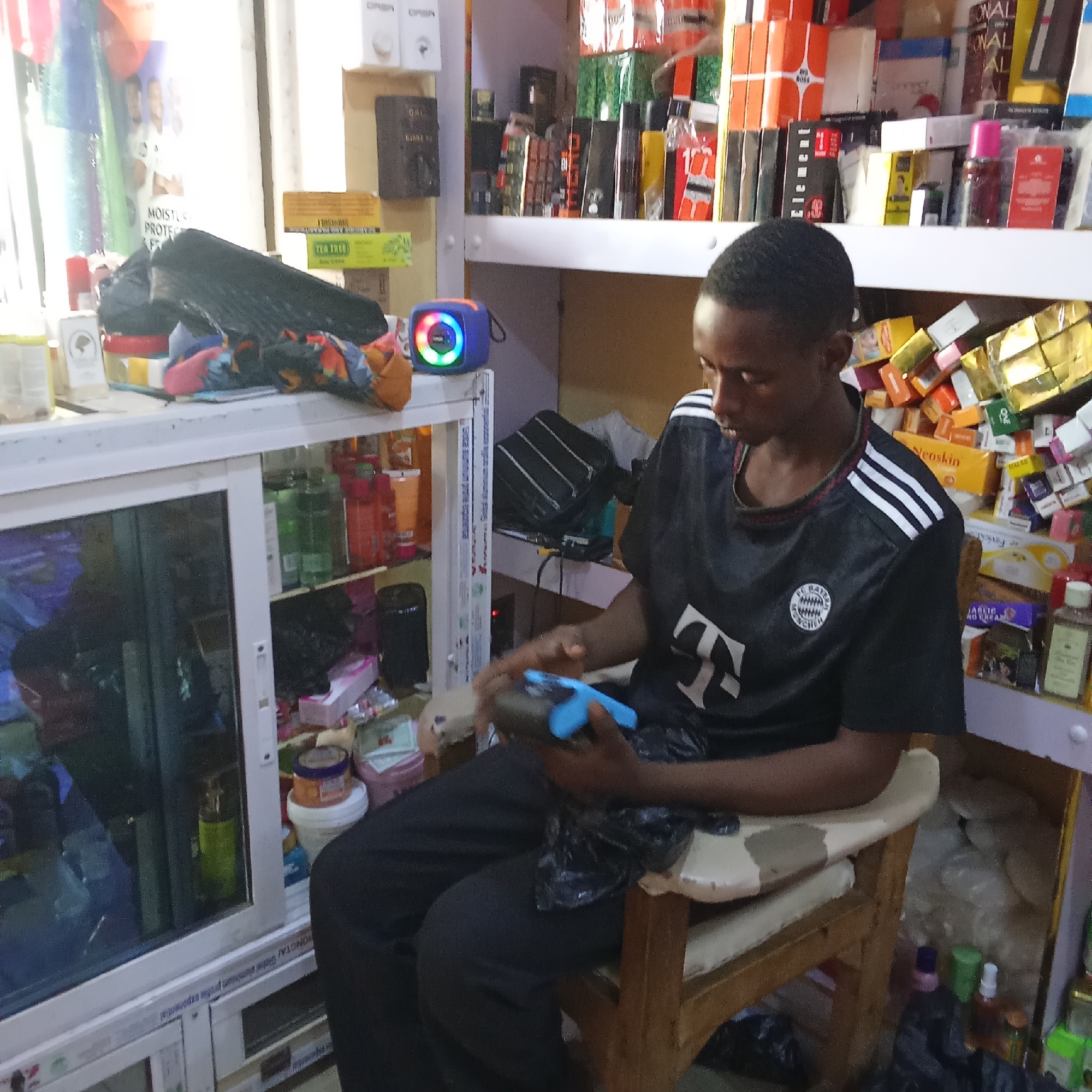 A young man sits in a retail shop in Akwanga, Nigeria, playing a video game on a blue controller. He is wearing a black Adidas shirt with white stripes on the sleeves and a Bayern Munich logo. The shop is filled with various products, including skincare items, perfumes, and other retail goods displayed on shelves and in a glass cabinet. A blue, light-up Bluetooth speaker sits on top of the cabinet.