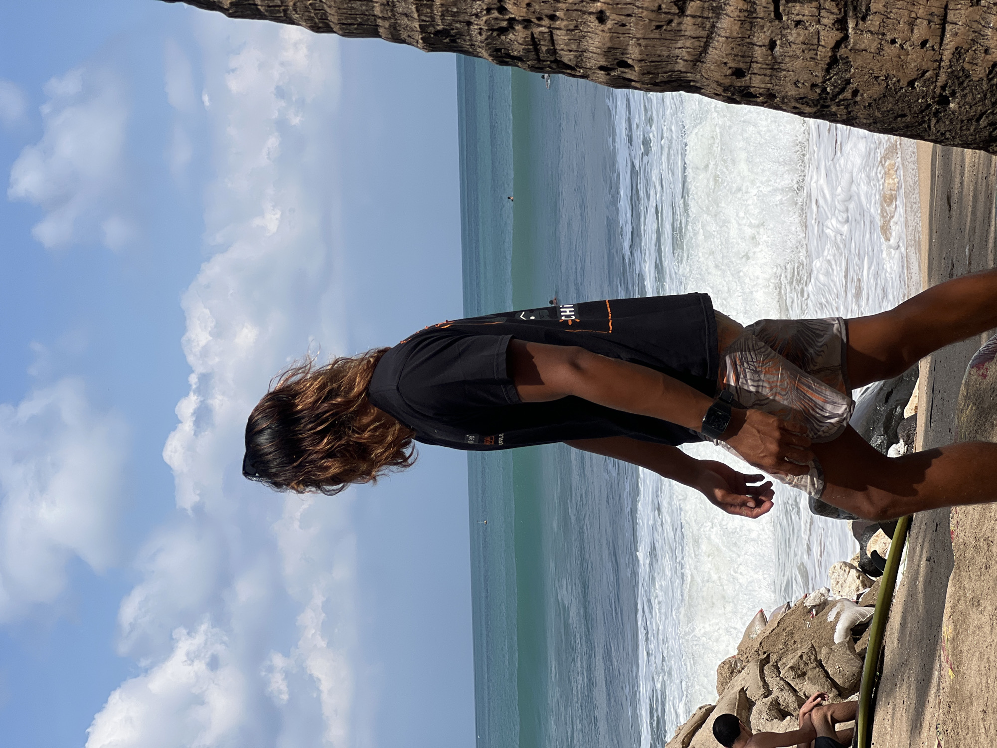 In Kuta, Indonesia, a young man with long, brown hair, wearing a black t-shirt and patterned shorts, stands on a sandy beach. He is facing away from the camera, looking out at the ocean, where waves are breaking. The sky is blue with scattered white clouds. To the right of the frame, a portion of a palm tree trunk is visible. In the foreground, to the left, are rocks and another person sitting. In the distance, several people are in the water, some appearing to be surfing. The overall scene is one of a sunny day at the beach.