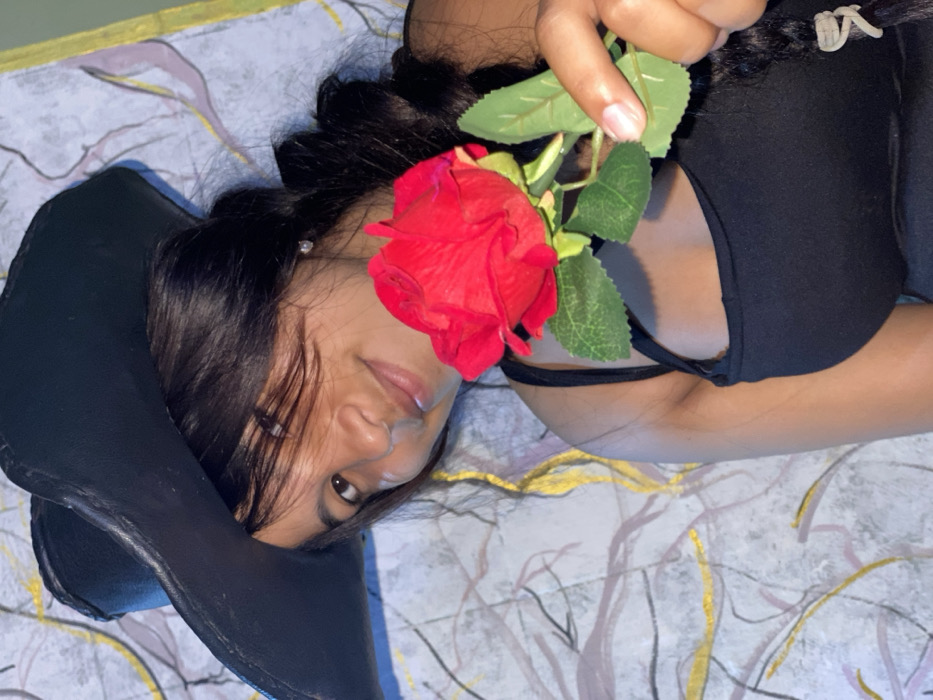 The image displays a young woman lying on a surface, viewed from a slightly elevated perspective, with the overall image rotated approximately 90 degrees clockwise. Her head is positioned towards the bottom-right of the frame, and her body extends diagonally upwards and to the left.
The woman has dark hair, likely black or dark brown, styled in what appears to be a braid or bun on the back of her head, with some loose strands framing her face. Her skin tone is tan. She is wearing a black top, possibly a tank top or camisole, with a thin strap visible over her right shoulder. A small, light-colored earring is visible on her left earlobe. Her left eye is open and looking towards the camera, while her right eye is partially obscured by a red rose. Her lips are slightly parted, revealing a neutral or soft expression.
She is holding a vibrant red rose in her right hand. The rose has several artificial-looking green leaves and is positioned in front of her face, partially covering her nose and right eye. Her fingers, with neatly manicured nails, are visible holding the stem of the rose.
To the left of her head, a dark, black object with a curved shape and subtle texture is present, possibly a wide-brimmed hat or a piece of folded dark fabric.
The woman is lying on a light-colored surface, which appears to be a blanket or sheet. This surface features an abstract, organic pattern composed of fluid lines and shapes in shades of grey, gold, and subtle purple or pink hues.
The lighting in the image is soft and even, casting no harsh shadows. The overall mood conveyed is relaxed and calm. No text is visible in the image.