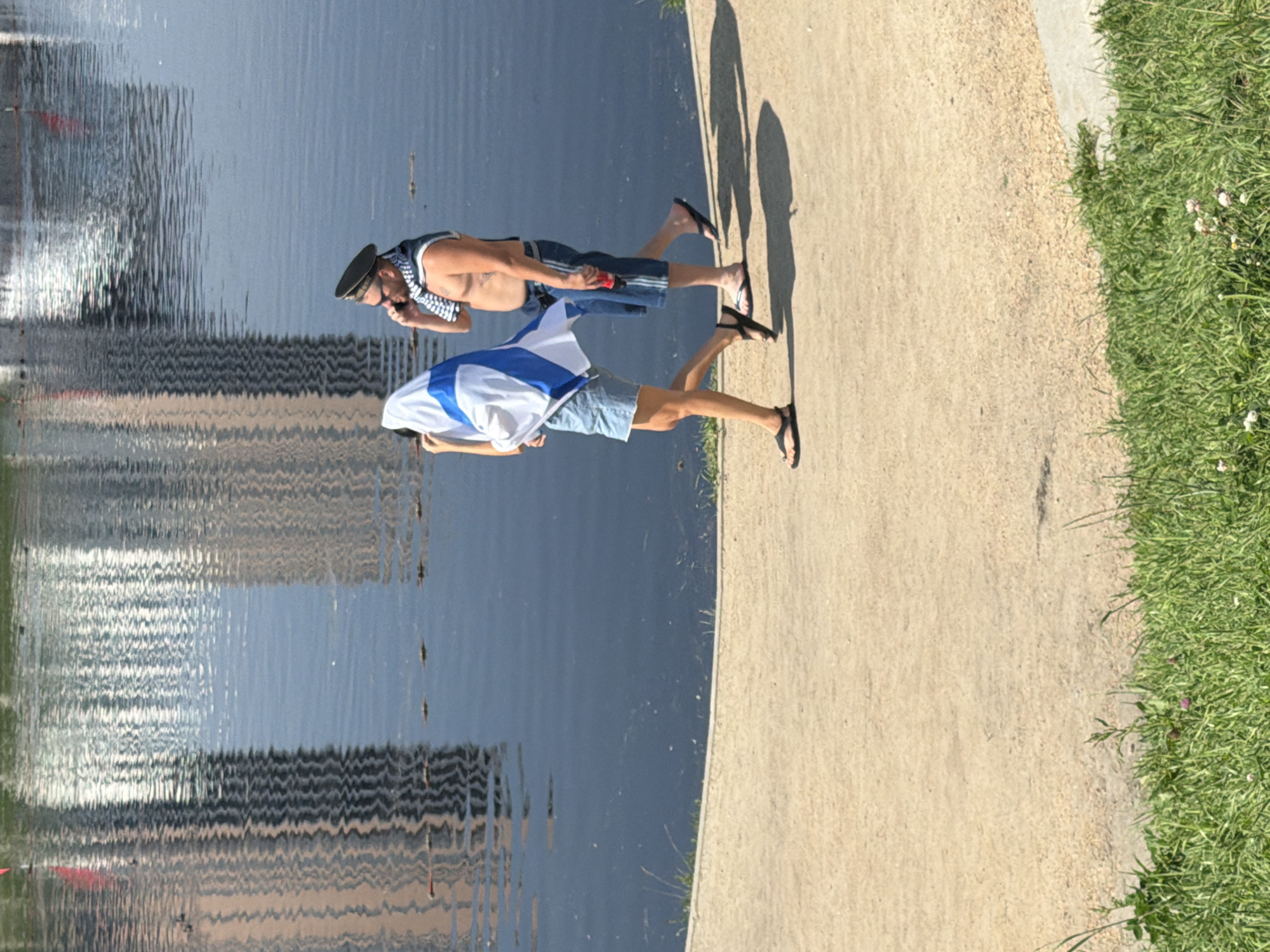 Two individuals are walking on a sandy path next to a body of water in Yekaterinburg, Russia. The person on the left is wearing denim shorts and is partially obscured by a white and blue flag. They are also wearing dark flip-flops. The person on the right is wearing denim shorts, a dark sailor's hat, sunglasses, and a scarf around their neck. They are holding a red can in their right hand and are on the phone. Buildings are reflected in the water, suggesting an urban environment. Green grass is visible in the foreground.