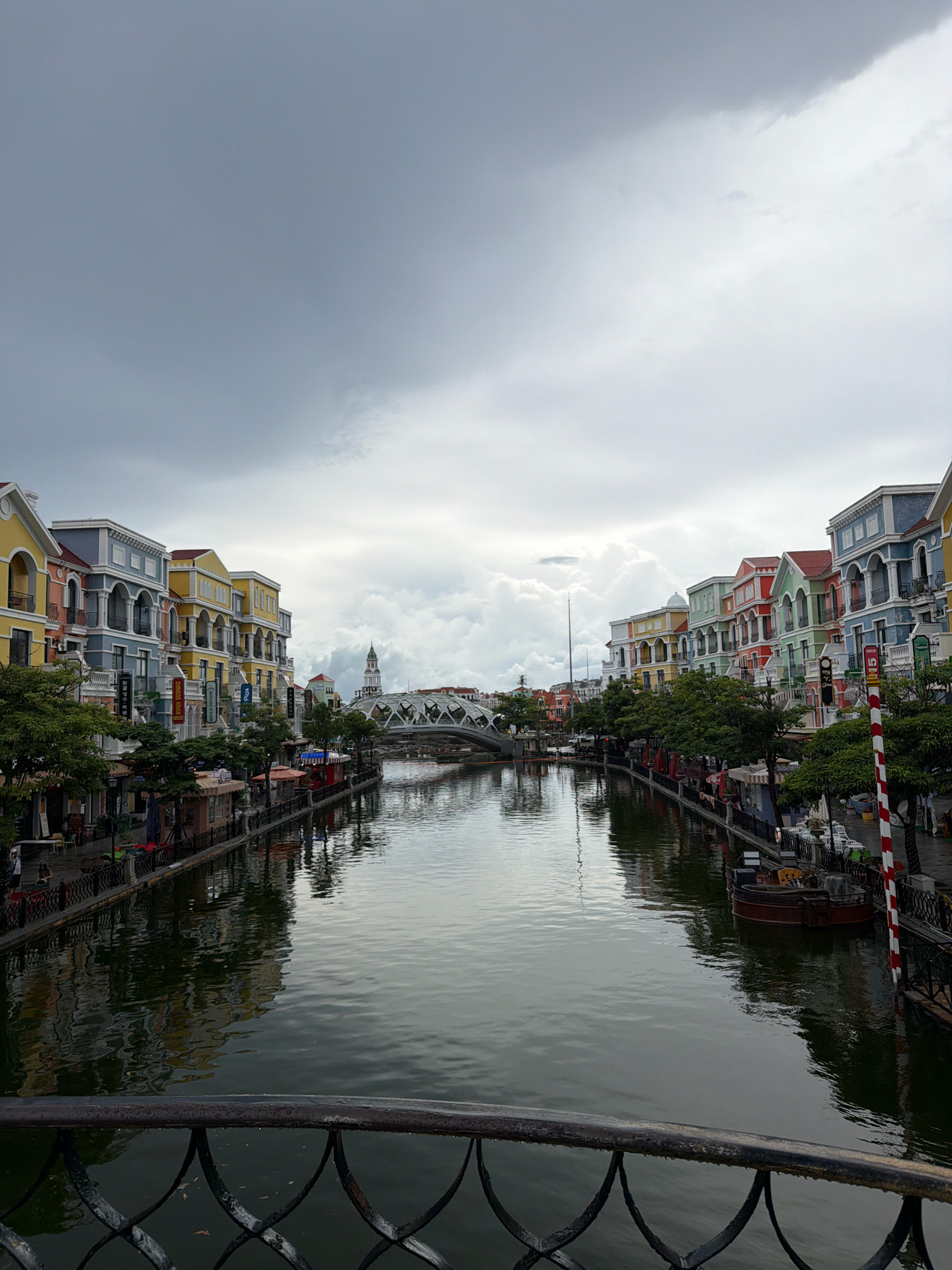 The image depicts a canal running through a European-style town on the island of Phú Quốc, Vietnam. Colorful buildings with classical architecture line both sides of the canal, featuring balconies, arches, and tiled roofs. Trees and outdoor seating areas are visible along the canal banks. A decorative bridge spans the canal in the distance, with a tower visible beyond it. The water in the canal reflects the buildings and the cloudy sky. There are a few boats docked along the sides and one small boat in the canal. The scene is calm and picturesque, with no people clearly visible in the foreground, but there are a few scattered figures on the sidewalks of the buildings. The sky is overcast, suggesting a cloudy day.