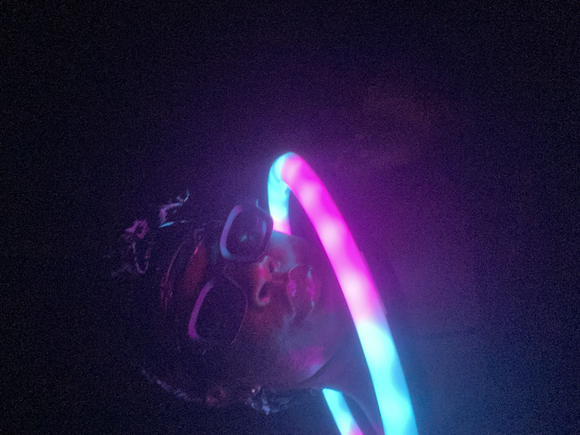The media file features a close-up, rotated shot of a person's face, partially obscured by vibrant neon lights, against a dark, grainy background. The primary subject is a person, possibly wearing sunglasses or goggles, with their face illuminated by a glowing ring. The ring displays a gradient of bright pink/magenta and cyan/blue light, creating a stark contrast with the dark surroundings.
The activity appears to involve the interaction of the person with this luminous object, possibly at a party, concert, or light art performance. The glowing ring seems to encircle or be positioned around the person's head, casting colorful reflections on their skin and eyewear. The image's composition suggests motion or a dynamic setting, emphasizing the light source.
Notable details include the extremely low light conditions, indicative of nighttime or an indoor environment where artificial light is dominant. The image has a pronounced digital grain or noise, typical of photography in dark settings. The facial features are softened by the intense, colorful light, making specific expressions hard to discern, though the overall mood is energetic and futuristic. No specific location cues for Maigizo, Nigeria, are visible within the frame, and there is no visible text.