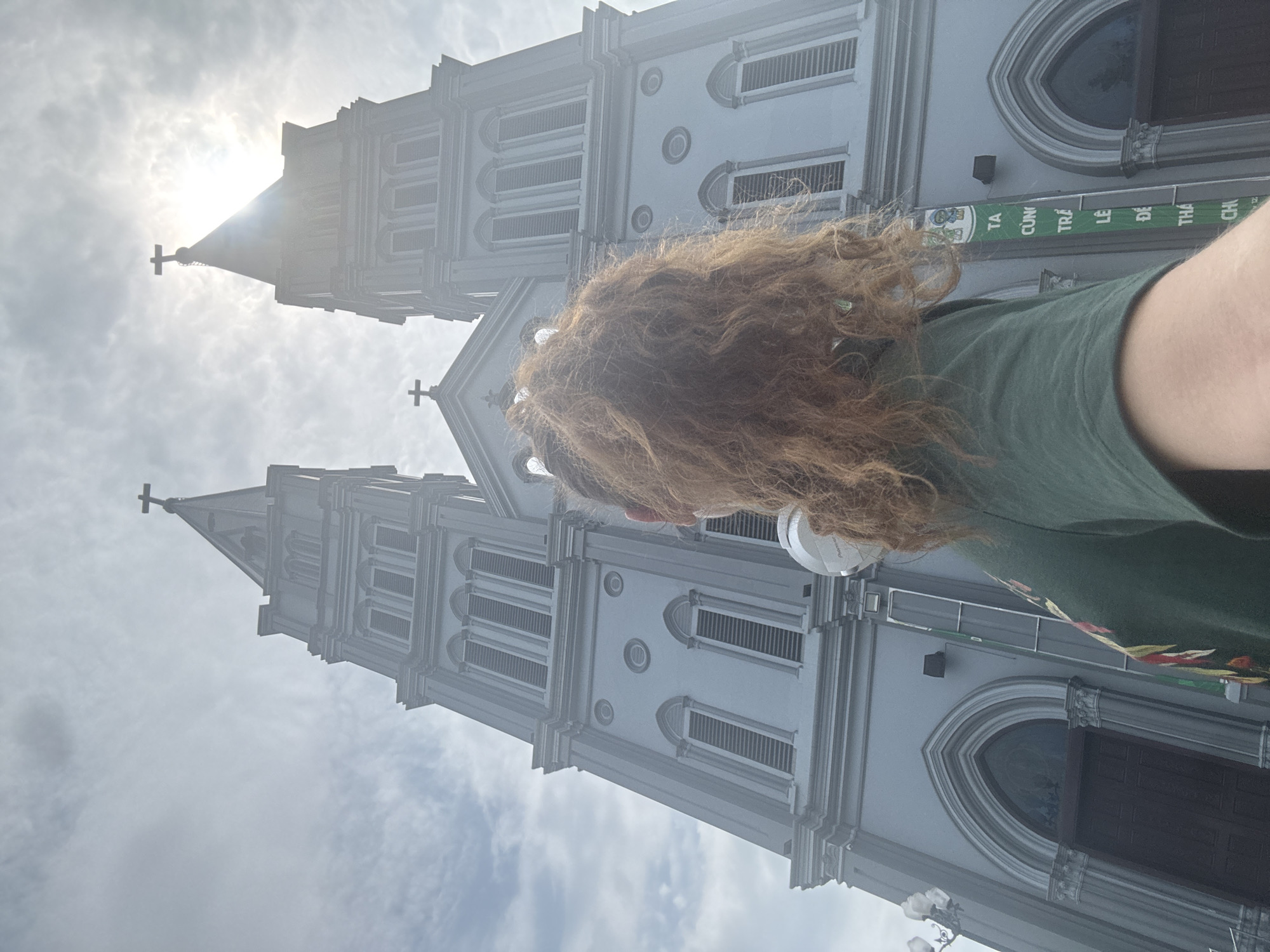 This is a first-person perspective shot of a person from the back, looking up at the imposing twin spires of St. Joseph's Cathedral, a prominent landmark in Hanoi, Vietnam. The cathedral, with its Gothic Revival architecture and two bell towers, dominates the frame. The sky is overcast with bright sunlight filtering through, creating a hazy, almost ethereal effect. The person has long, curly red hair and is wearing a dark green t-shirt with a colorful floral or patterned design on the left sleeve. They are holding a white device, possibly a portable speaker, in their right hand. A vertical banner with Vietnamese text is visible on the right side of the cathedral's facade. The image captures a moment of awe and contemplation of the historic architecture.