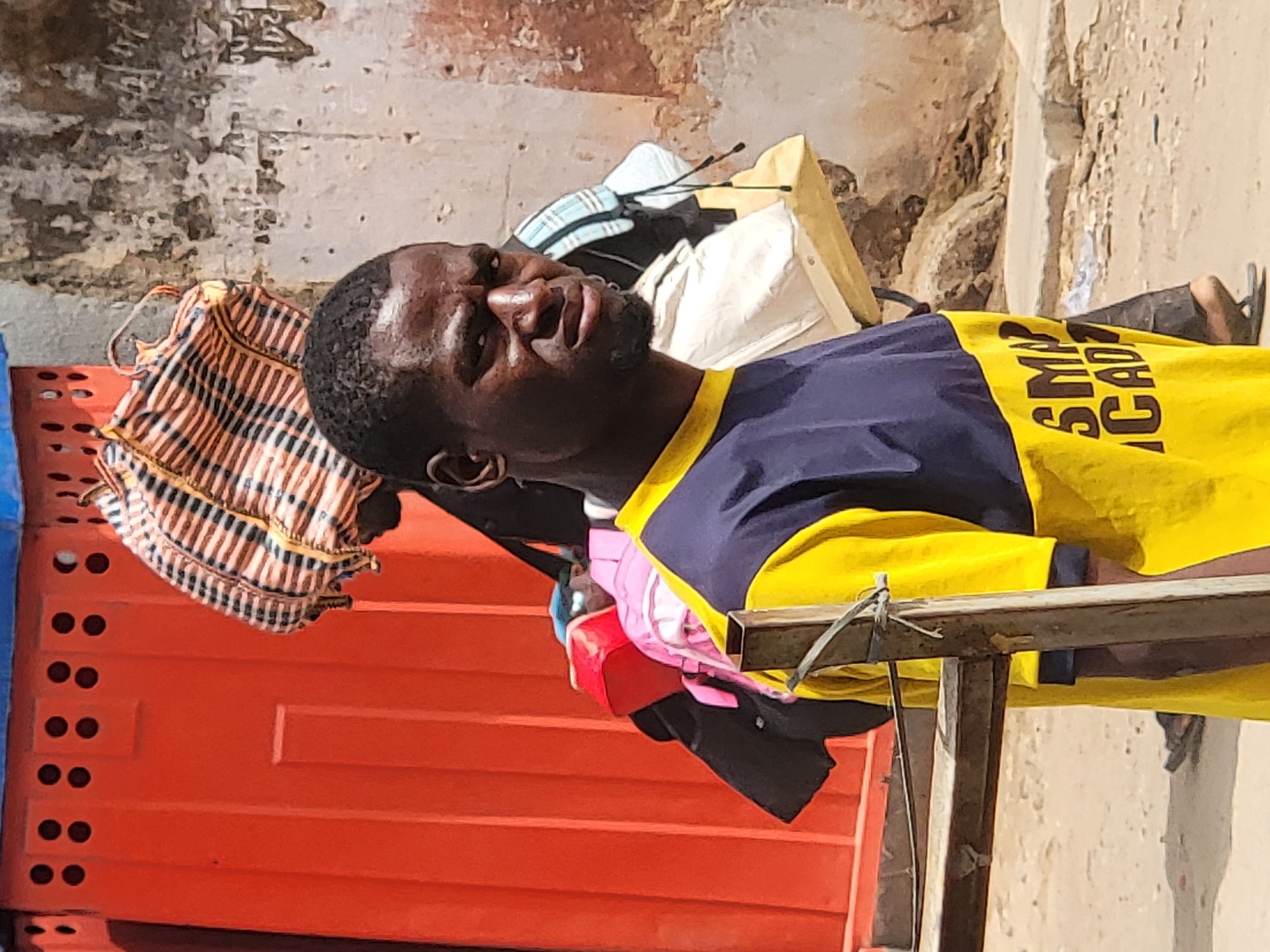 An adult Black male is depicted in a brightly lit outdoor setting in Maiburiji, Nigeria, under clear, sunny conditions. He is framed from the chest up, positioned slightly to the right of the center, and looks upwards and to his right with a neutral to contemplative expression. He has short, dark hair and a light beard or stubble.
He is wearing a short-sleeved t-shirt that is split into two colors: navy blue on the upper portion and bright yellow on the lower part and sleeves. Black text, "SMA ACAD", is visible on the yellow section of his shirt, likely part of "SMA ACADEMY".
The background features a weathered, light-colored wall with patches of darker discoloration. To the man's left, a vibrant orange, ridged structure, possibly a plastic crate, barrier, or part of a stall, occupies the left side of the frame. A striped bag in earthy tones of brown, orange, and white rests on or near this orange structure. Behind his head, a light blue and white striped bag or backpack, along with other light-colored fabric items, are visible.
A wooden beam or frame, secured with what appears to be wire or twine, extends horizontally across the lower right foreground, partially obscuring parts of the scene. Beneath the man's right arm and near the wooden frame, various small objects, including a red container, are scattered. The overall impression is an active outdoor environment, possibly a market or street scene.