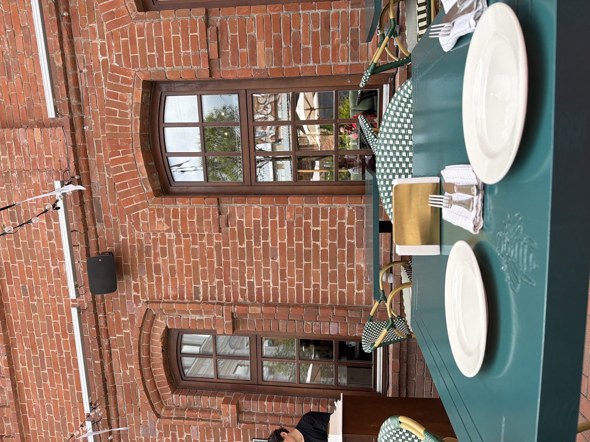 The image depicts an outdoor seating area of a restaurant with a brick facade and large arched windows. A dark teal table in the foreground is set with two white plates, silverware, and napkins. To the left, a person wearing a black shirt is partially visible behind a counter or serving station. Green and white checkered chairs are placed around other tables, some of which are visible in the background, reflecting the exterior scenery. String lights and a speaker are mounted on the brick wall above. The overall setting suggests a dining establishment in Ufa, Russia, based on the context provided.