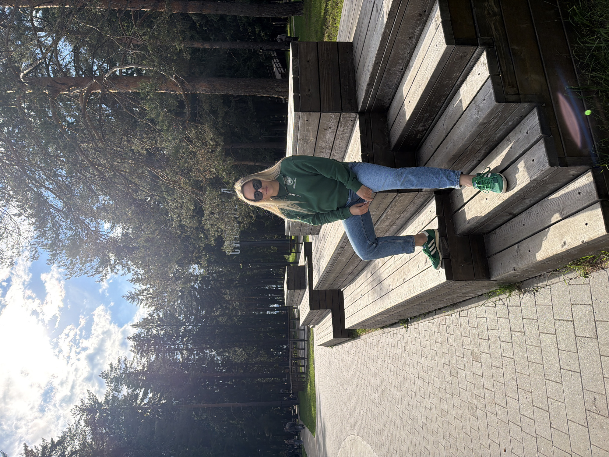 A young, blonde woman is perched on a wooden structure of steps outdoors in Votkinsk, Russia. She is wearing sunglasses, a green sweater, blue jeans, and green Adidas sneakers. The wooden structure appears to be tiered seating. A paved area is adjacent to the steps. A dense forest stands in the background. The sky is partly cloudy.