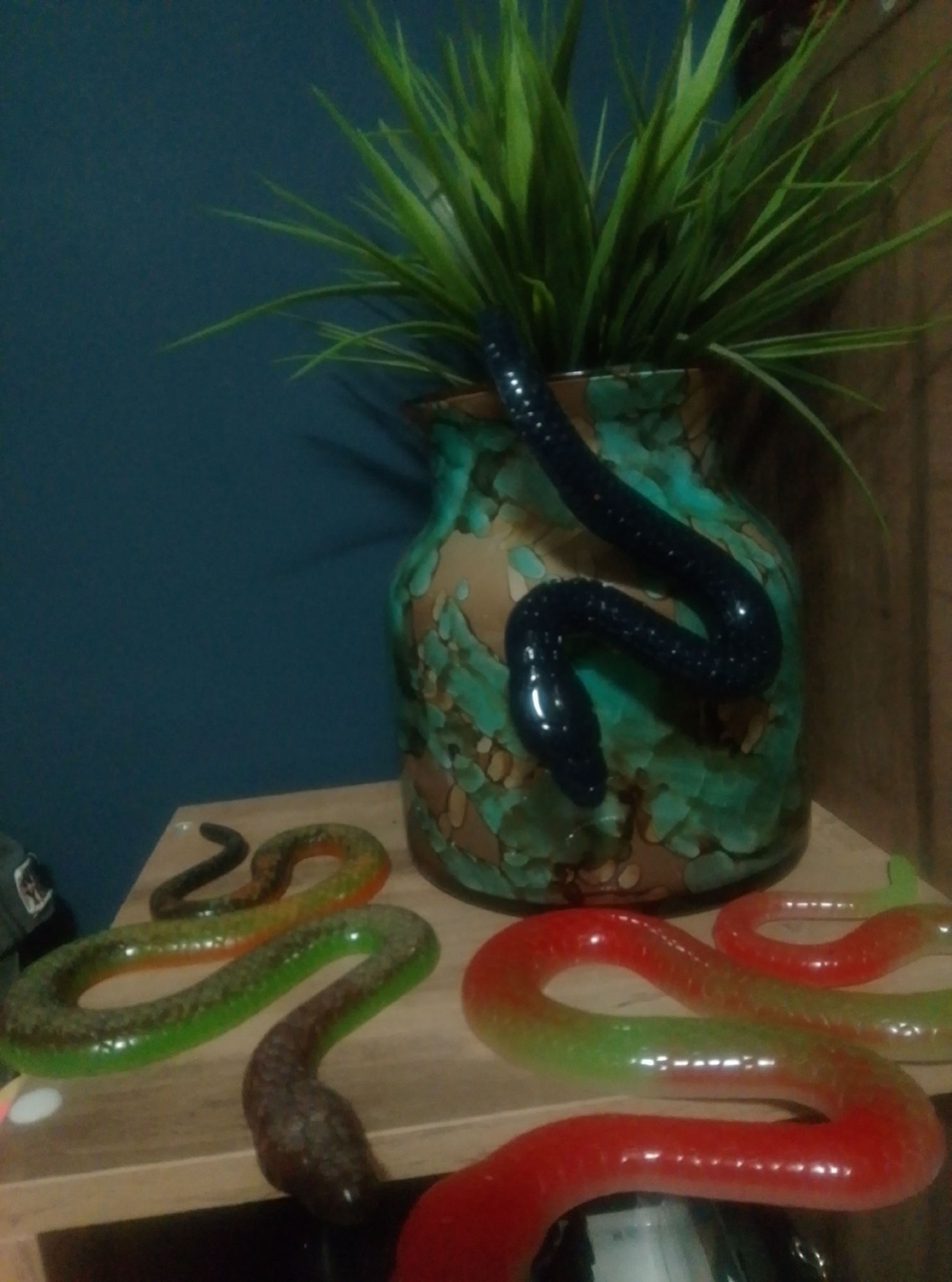 This is a medium-shot, indoor, eye-level, horizontal photograph of decorative, snake-shaped objects on a wooden surface in front of a teal-colored wall. The objects appear to be made of a translucent, pliable material, possibly candy or plastic. One black snake is coiled around a decorative vase containing green artificial plants. Two other snakes are on the wooden surface in the foreground: one is green and orange, and the other is red and green. The vase is green and brown with an irregular pattern. The lighting in the image is dim, and the background is blurred. The scene is likely a home or retail environment.