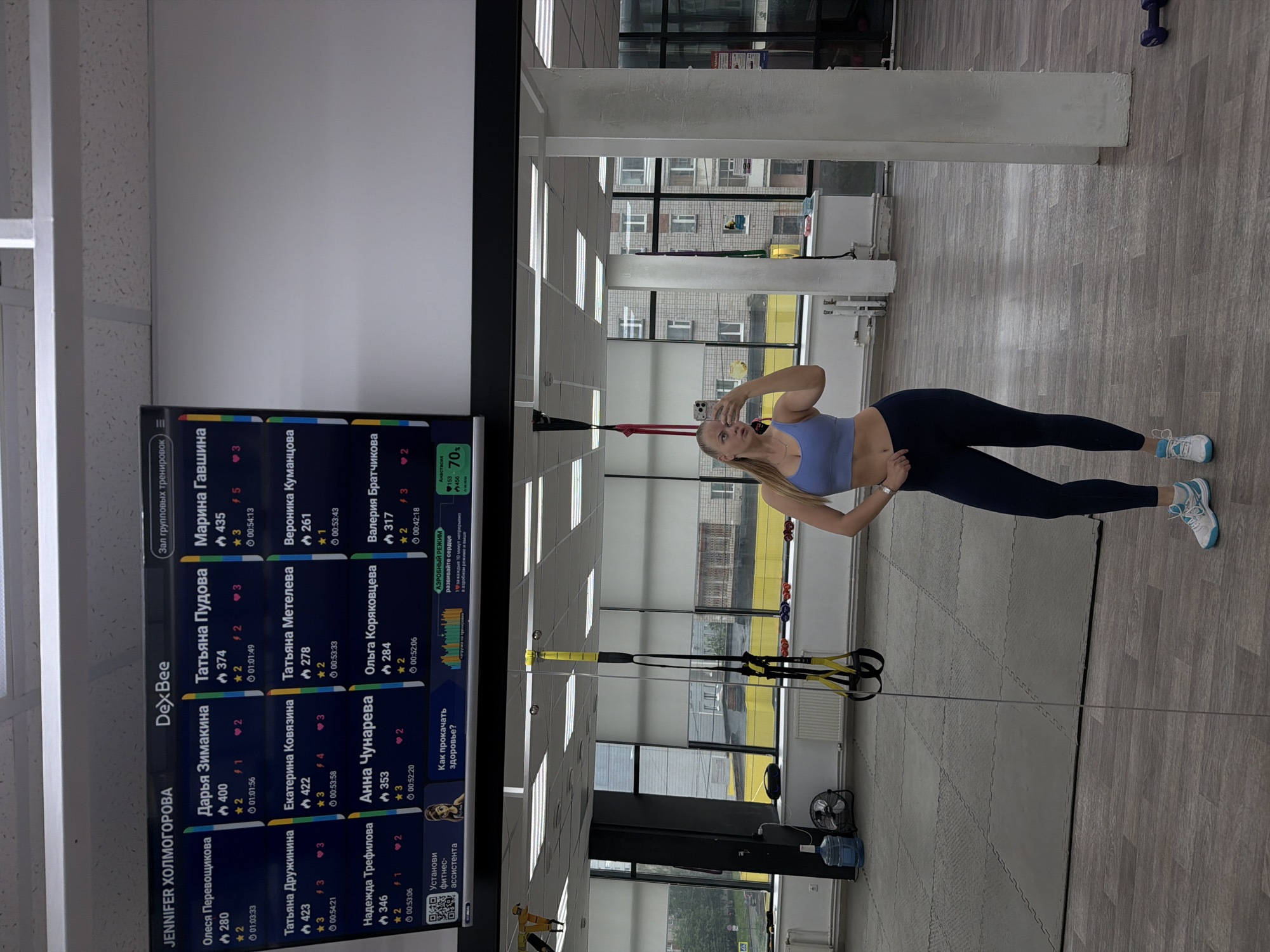 The image shows a woman taking a mirror selfie in a gym in Izhevsk, Russia. The woman is wearing a light purple sports bra and dark blue leggings, and blue and white sneakers. She is holding a phone in her right hand. A display screen with performance statistics of other gym members is visible on the left. Gym equipment, including TRX suspension straps, is present in the background. Through the windows, one can see a cityscape. A blue dumbbell is visible in the background of the gym. The gym has rubber mats and a fan, along with a water bottle.