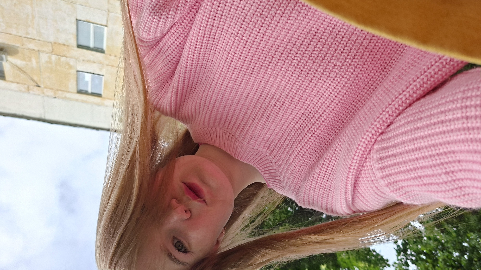 A low-angle, upward-looking photograph captures a young woman with long, light-colored hair and fair skin, likely taken as a selfie from below. The image is rotated, presenting the woman's face towards the left side, looking downwards into the lens. She wears a prominent, chunky pink knitted sweater, which fills a significant portion of the frame. Her eyes are light-colored, and she has pink lipstick on her lips, with a soft, neutral expression.
In the background, to the left of the woman's head, is a multi-story building with light-colored, possibly beige or yellow, walls and several rectangular windows. Above the building, a bright, partially cloudy sky is visible. Towards the bottom right of the frame, green foliage from trees or bushes suggests an outdoor setting in Chaikovsky, Russia. A small, indistinct yellow or orange element is visible at the very top right edge, possibly part of a ceiling or another object in the immediate foreground. The time of day appears to be bright daylight. There is no visible text.