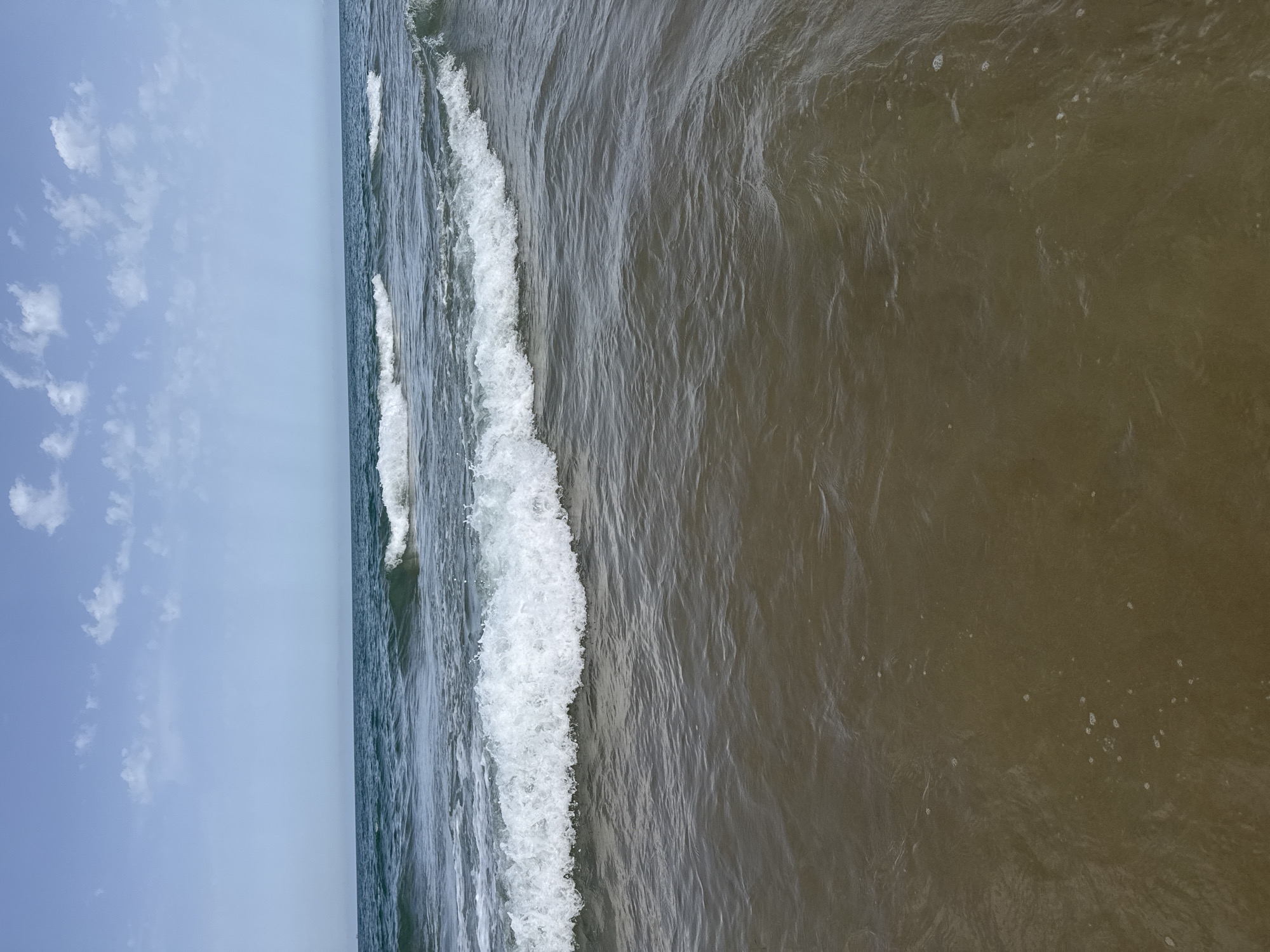 The image depicts a serene coastal scene at Gandia, Spain, under a partly cloudy sky. The foreground is dominated by the shallow, murky waters of the sea, with small bubbles and ripples visible on the surface. Gentle waves, capped with white foam, are breaking and rolling towards the shore. In the distance, the horizon line separates the sea from a clear, pale blue sky dotted with scattered white clouds. There are no people or man-made structures visible in this snapshot of the Gandia coastline.