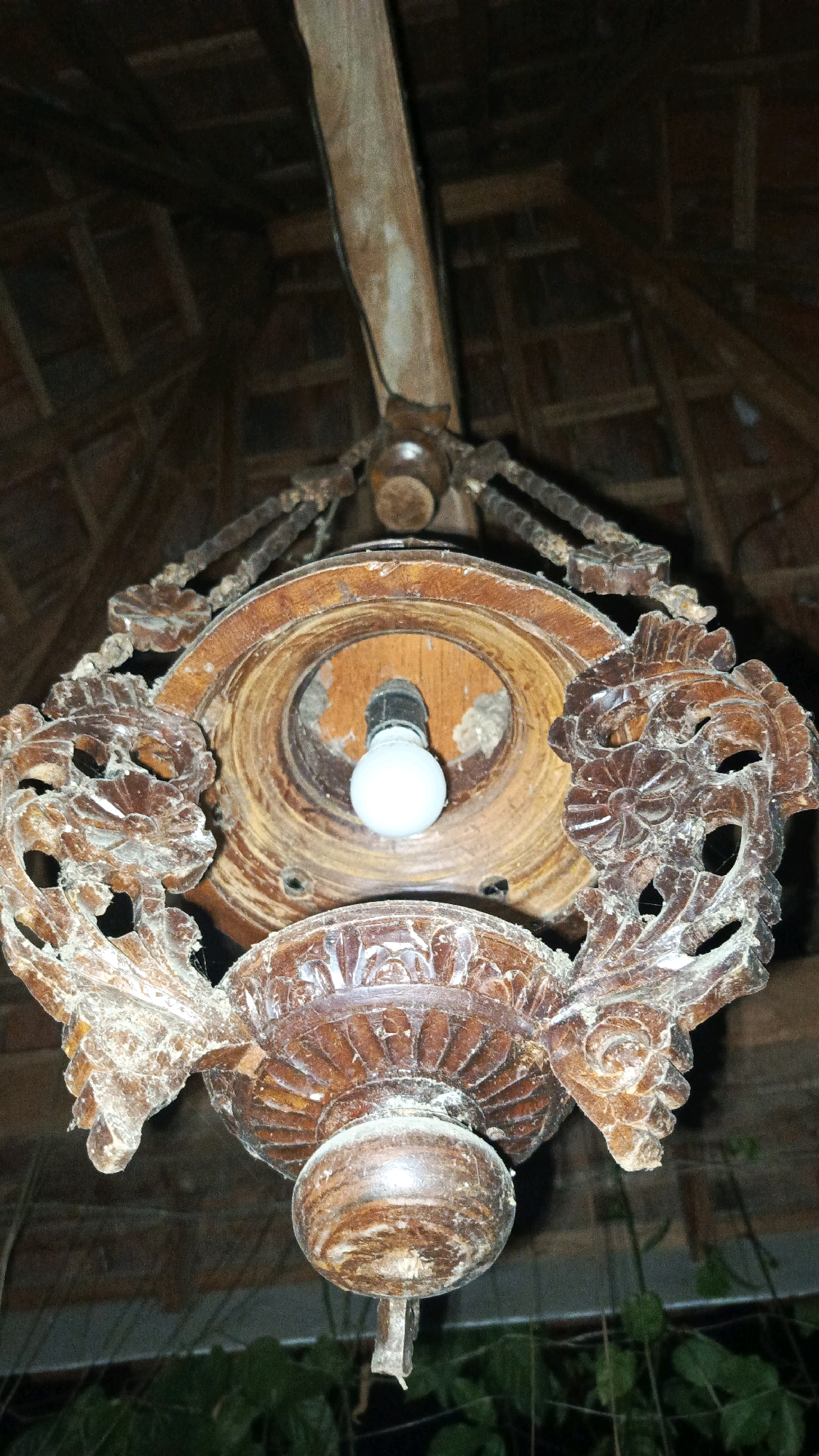 This is a close-up, low-angle shot of an old, ornate wooden chandelier hanging from a wooden beam and thatched roof. The chandelier is intricately carved with floral and scrollwork motifs, and appears to be aged and dusty, with some areas of wear and tear. A single, bare light bulb is visible in the center of the fixture. The background shows the texture of the thatched roof and wooden structural elements. The lower part of the image shows blurred green foliage, suggesting it is outdoors or in a semi-enclosed space with plants. The overall impression is one of rustic charm and antiquity. This scene is located in Babadan, Indonesia.