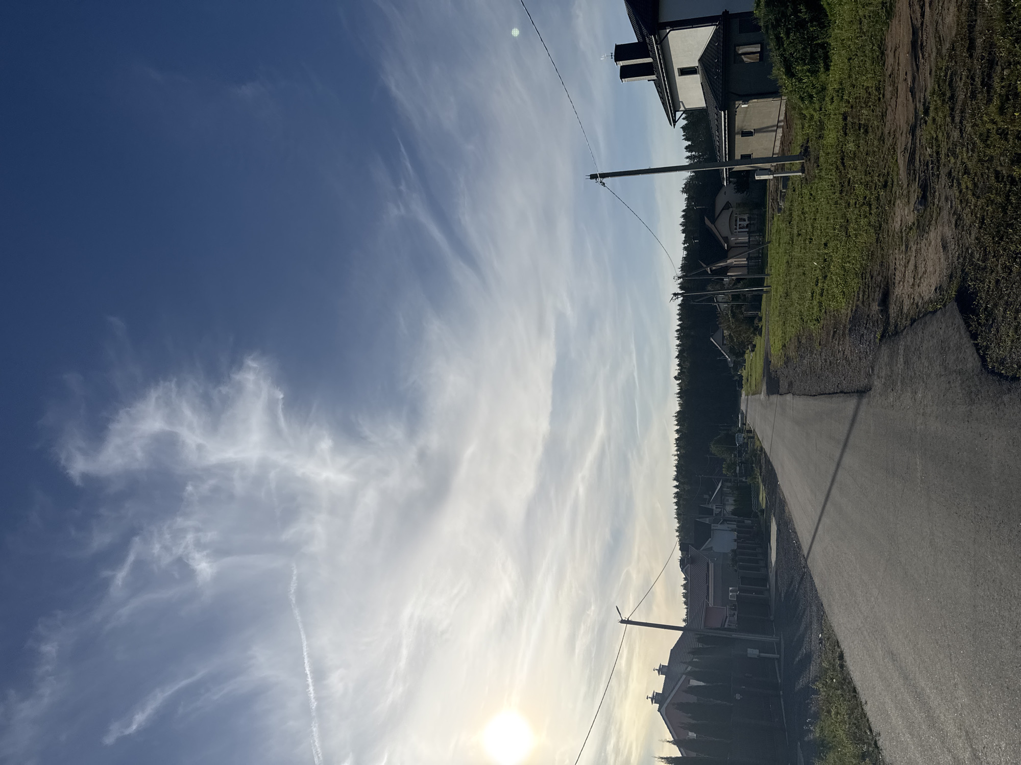 The image shows a paved road in a suburban area, likely within the Бараўлянскі сельскі Савет of Belarus. On the left side of the road, there are several houses with fences and manicured hedges. The houses have pitched roofs and are of varying architectural styles. Further down the road, a dense forest forms the background. On the right side of the road, there are more houses, including a prominent white building with a black roof and a smaller adjacent structure. Power lines and utility poles are visible throughout the scene, running along the road. The sky is blue with wispy white clouds, and the sun is setting on the left side, casting long shadows. The overall impression is of a quiet, residential neighborhood.