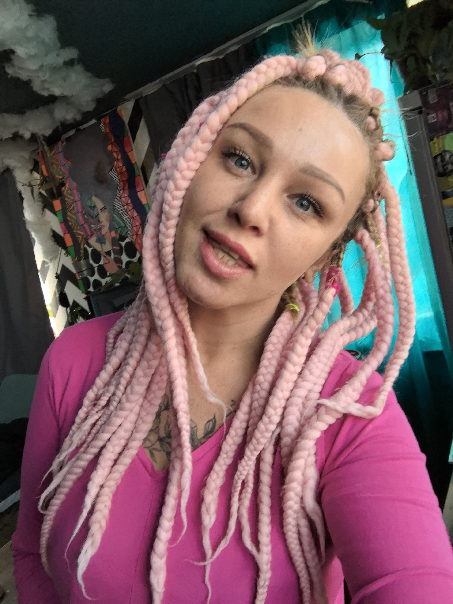 A woman with pink braided hair wearing a pink long-sleeved shirt is in an indoor setting. Behind her is a wall with a blue curtain partially visible on the right, and a colorful patterned tapestry to the left with what appears to be a portrait of a person on it. There are also some white, cloud-like decorations hanging near the tapestry. There is a tattoo on her chest visible just below her neck. The exact location in the city of Noviy, Russia cannot be determined from the image alone.