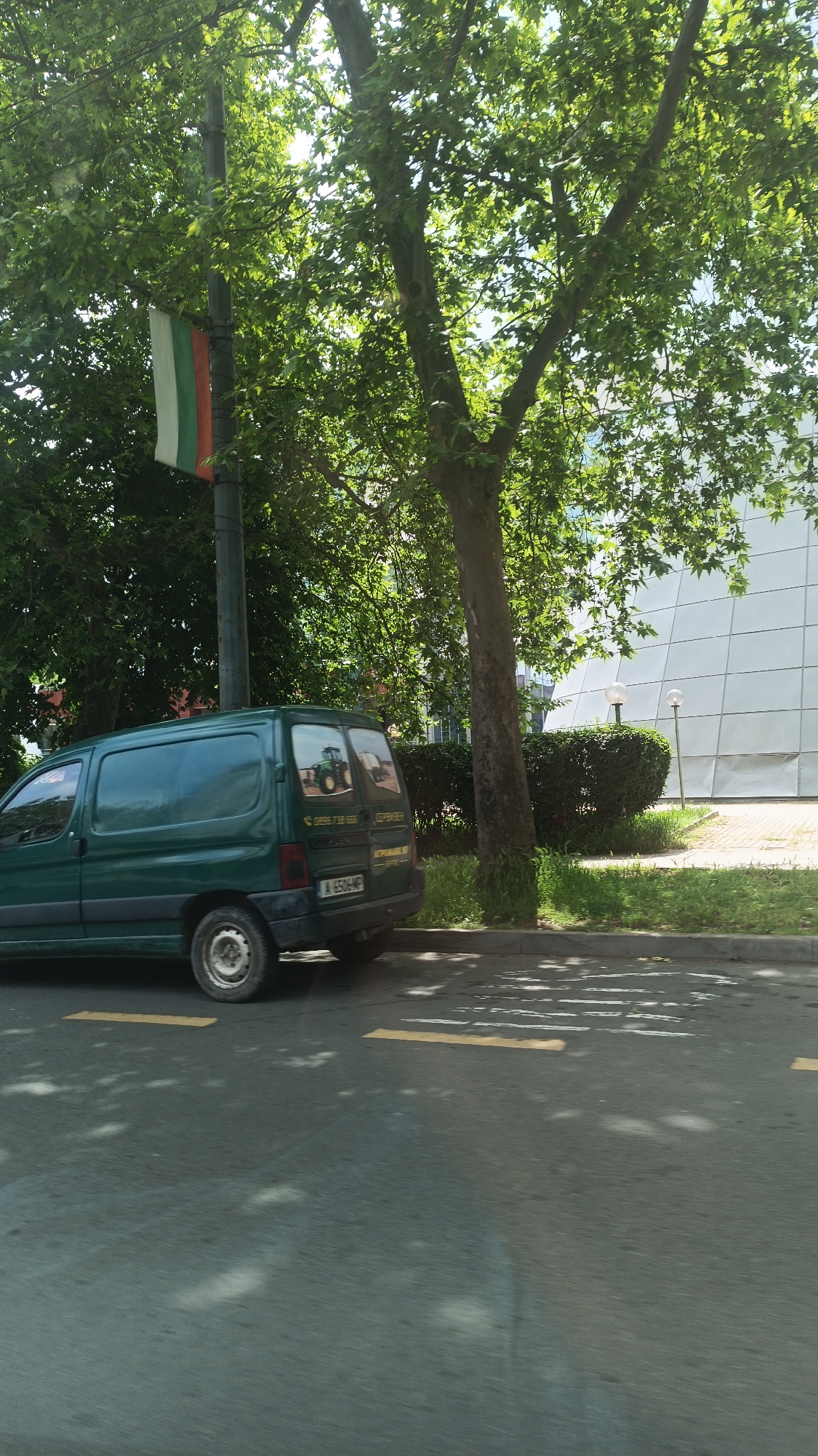 A daytime shot shows a green Peugeot Partner van parked on the right side of a street in Burgas, Bulgaria. The van has agricultural-themed images on its rear windows. A pole displaying the Bulgarian flag is visible next to the van. A tree is next to the van, casting shadows on the road. In the background is a building with a distinct, tiled, white facade and manicured greenery with lampposts. The road has yellow lane markings.