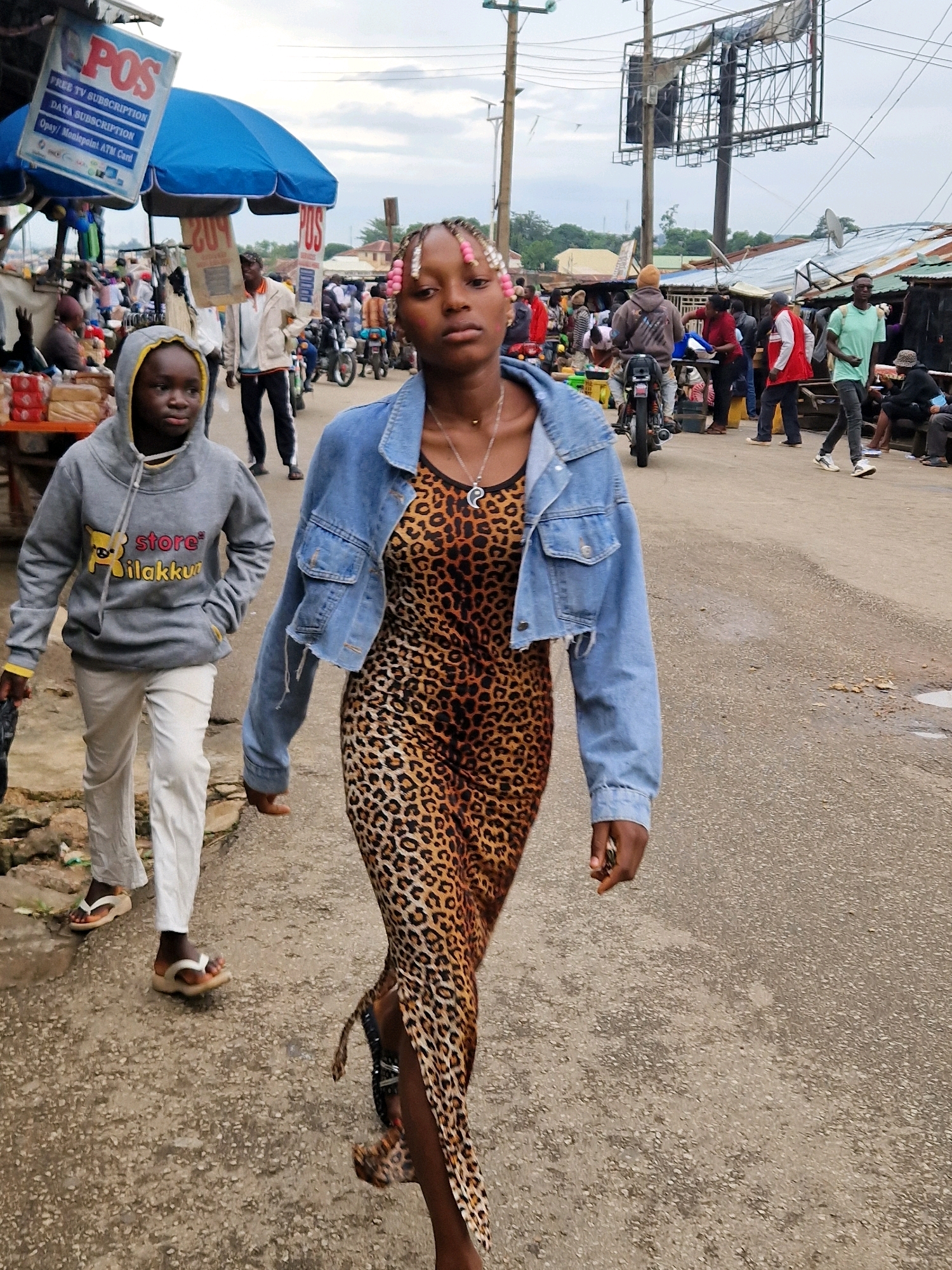 In an outdoor street market scene, two individuals are walking towards the camera. The primary subject is a young Black woman in the foreground, wearing a denim jacket over a leopard print dress and a necklace. Her hair is styled with pink beads. To her left and slightly behind her, a younger Black person in a grey hooded sweatshirt and white pants walks ahead. The background is bustling with activity: other people are visible, some standing and some on motorcycles, amidst market stalls and makeshift structures with corrugated metal roofs. A blue umbrella and a large billboard structure are also present in the scene. The ground is paved with rough asphalt. The overall impression is a vibrant and busy urban marketplace.