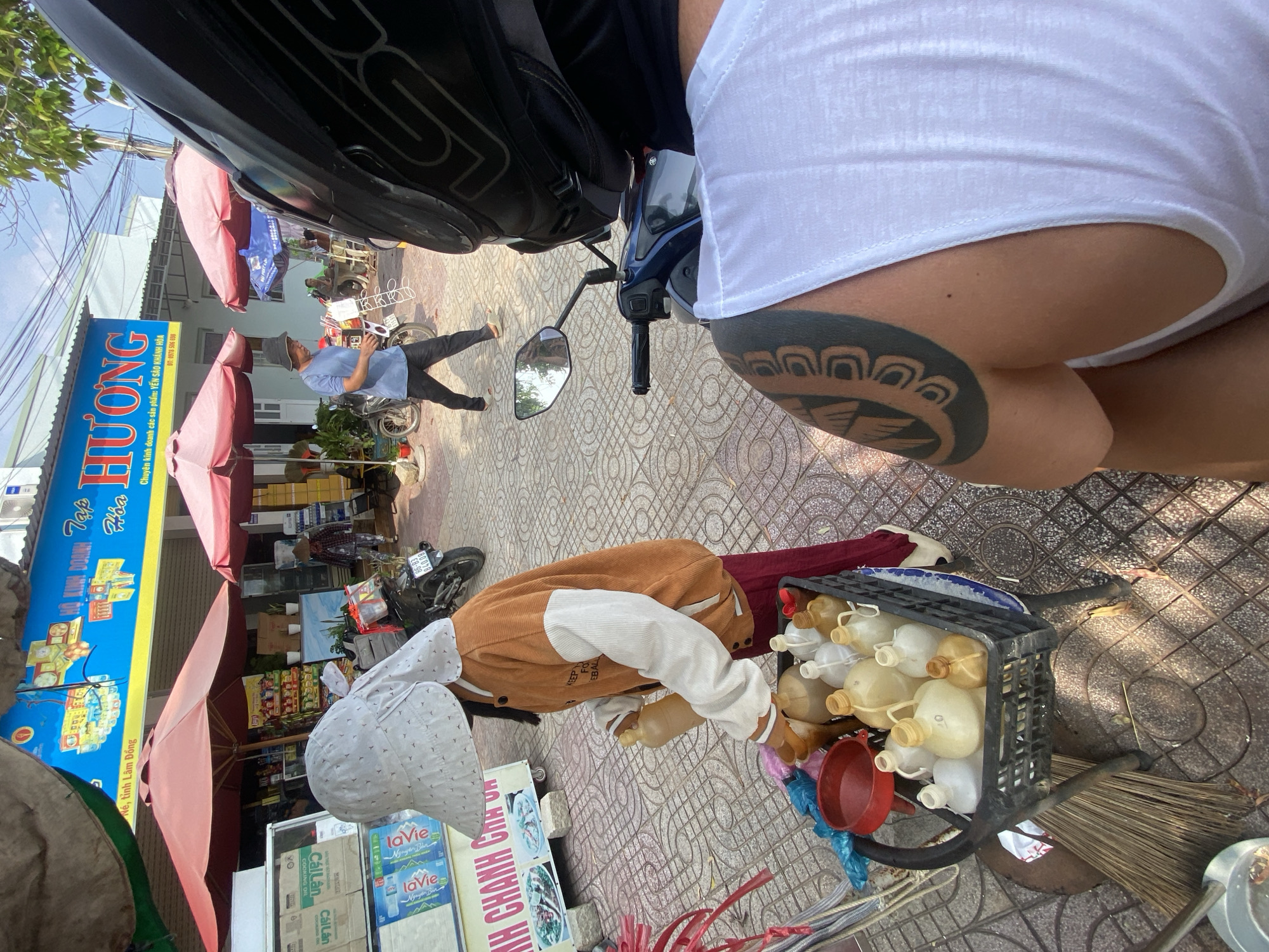 The image depicts a street scene in Mũi Né, Vietnam, likely in front of a small shop or market. In the foreground, a person wearing a white sun hat and an orange and white jacket is holding a brown plastic bottle over a black crate filled with various plastic containers. Beside the crate, a red plastic funnel and a broom are visible. To the right, the shoulder and back of another person wearing a white tank top and a black motorcycle helmet are in view, showcasing a distinctive tattoo on their arm. In the background, a blue sign with yellow lettering reads "TẬP HÓA" and "HƯƠNG," indicating a general store. Several red umbrellas are set up in front of the store, providing shade. People are walking in the background, and motorcycles are parked nearby. The ground is paved with patterned tiles. The overall environment suggests a commercial area in a sunny, urban setting.