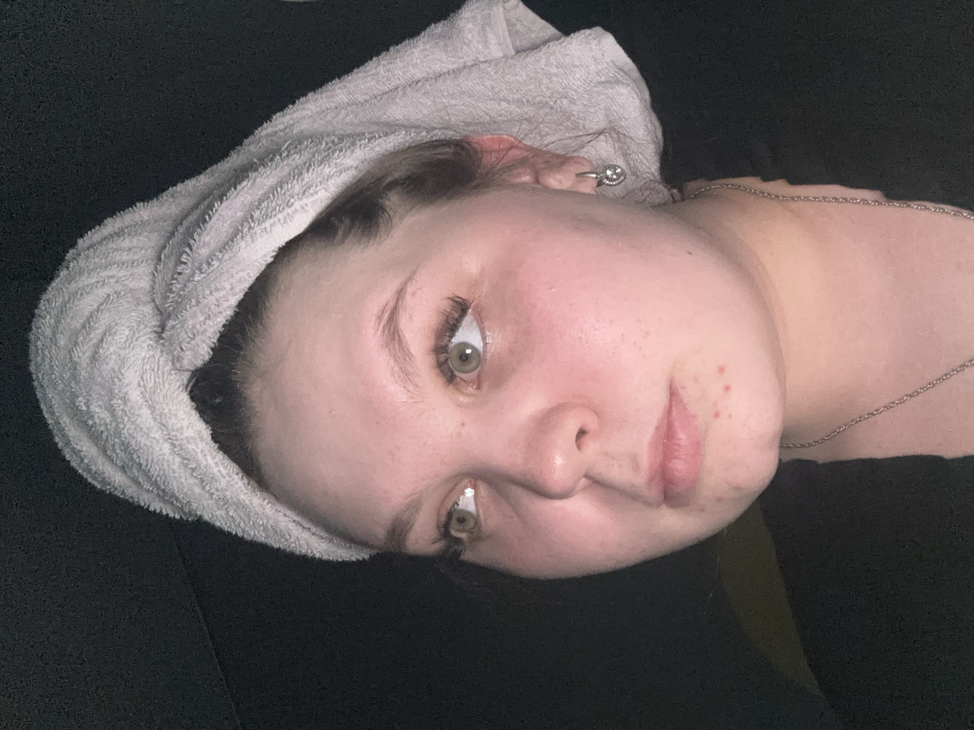 The image captures a close-up, chest-up portrait of a young adult female, positioned to the right of the frame, with her head turned slightly to her left. She wears a light gray or white terry cloth towel wrapped around her head, obscuring most of her dark hair, with strands visible near her temples. Her fair complexion shows areas of redness on her cheeks and under her eyes, and small, red blemishes are present on her chin and right cheek. Her light-colored (green/hazel) eyes are directed upwards and to the viewer's left. Prominent eyelashes are visible. A silver-colored hoop earring, featuring a round, decorative element, is affixed to her right earlobe. A thin, silver-colored chain necklace is visible around her neck, resting against a black garment worn on her upper body. The background is uniformly dark and featureless, providing no specific contextual elements. The location of the scene is Chaikovsky, Russia.