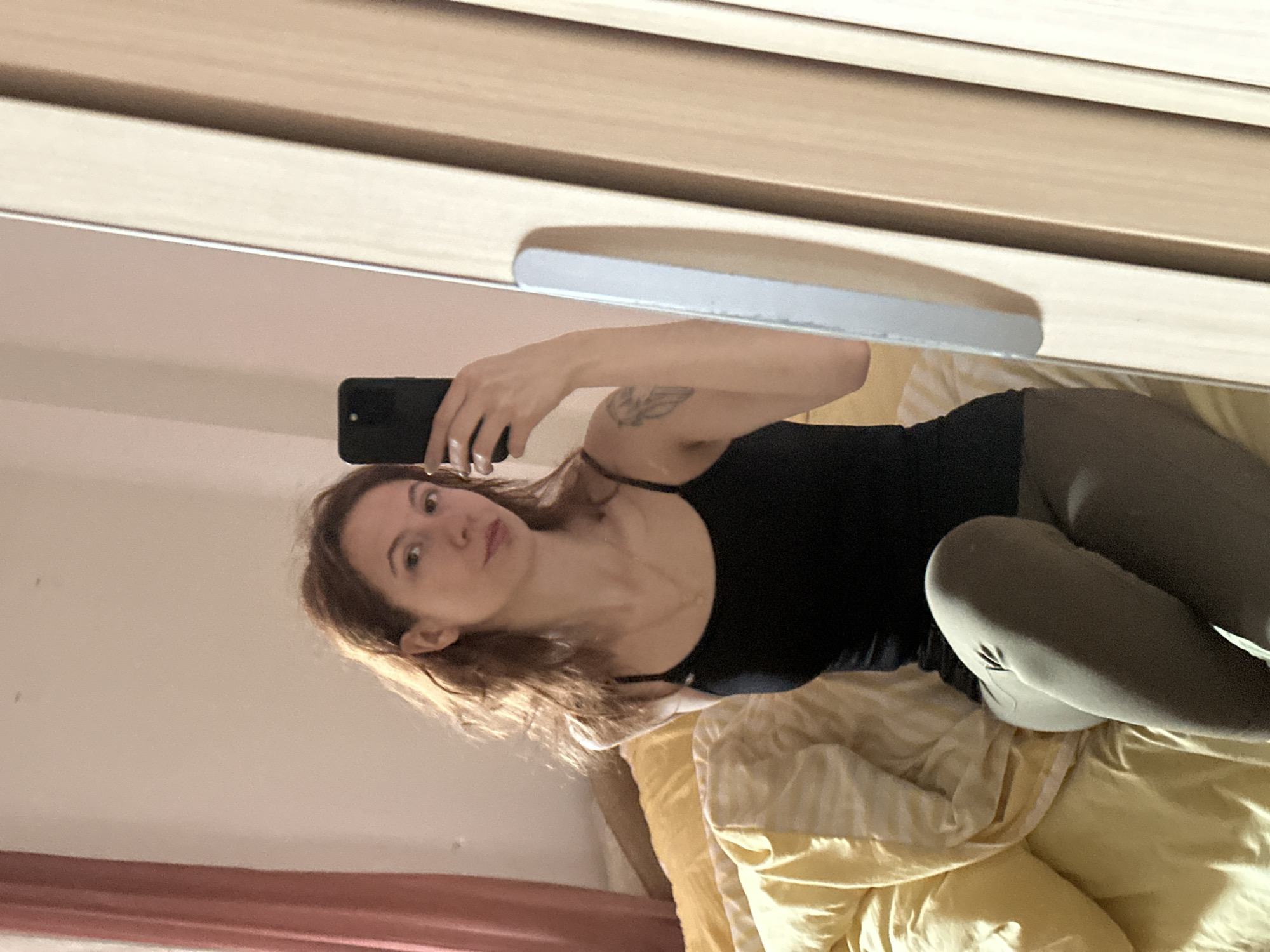 A young woman with red hair is taking a selfie in a mirror. She is wearing a black tank top and grey leggings. She is lying on a bed with a yellow comforter. The bed is in a room with beige walls and a wooden dresser. It is not possible to determine the location in the city of Novi Sad, Serbia.