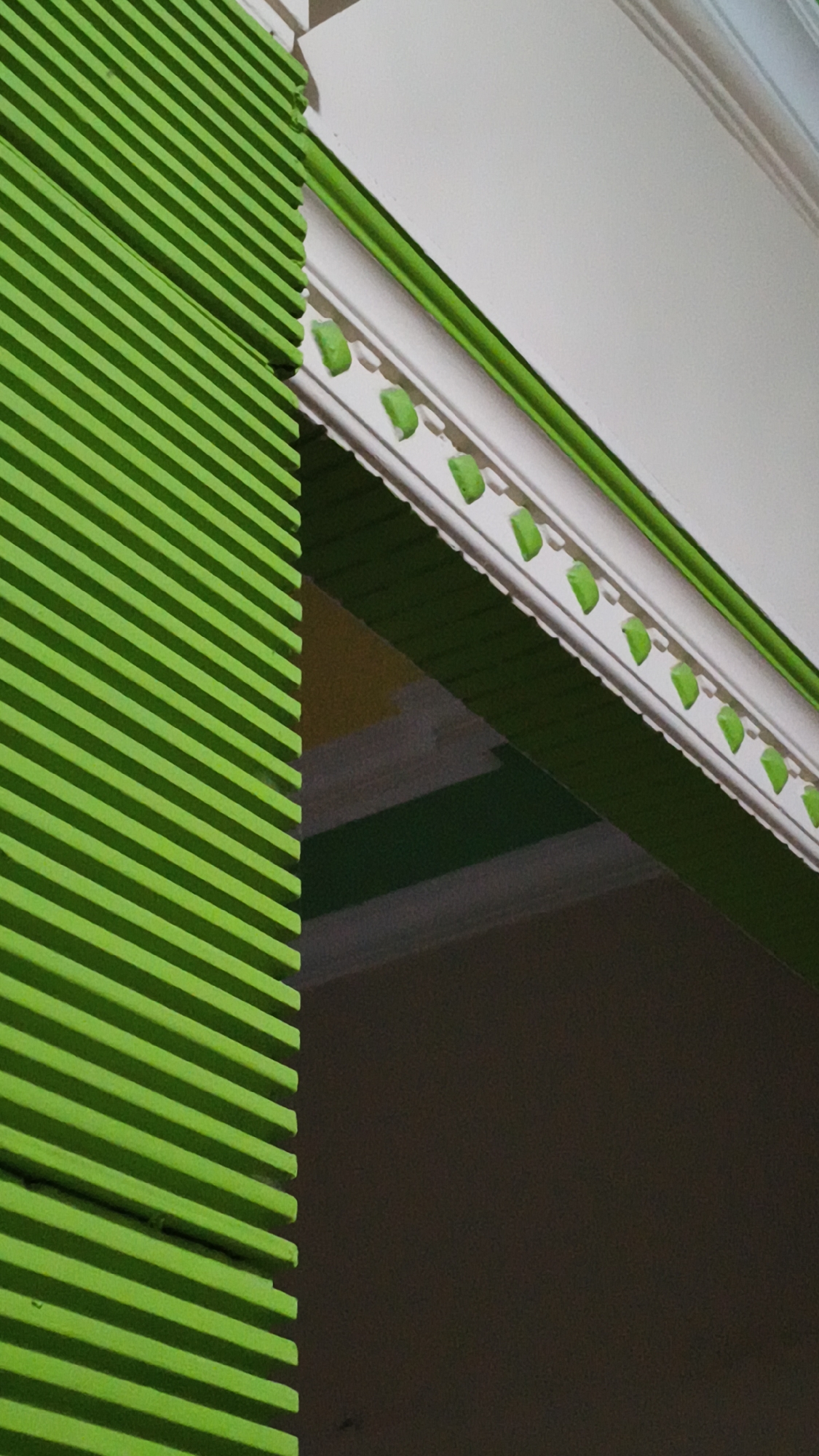 The image is a close-up, abstract shot of architectural details, likely of a building in Kerep Kidul, Indonesia. On the left, a bright green slatted structure dominates the frame, its parallel lines running diagonally from bottom left to top right. To the right of the green structure, a white, ornately decorated cornice runs horizontally across the upper portion of the image. This cornice has a series of small, green, leaf-like embellishments spaced evenly along its lower edge. A thin, bright green band runs along the upper edge of the cornice. Below the cornice, a dark, shadowed area fills the lower right and extends to the right edge of the frame. In the middle ground, between the green slats and the cornice, there are glimpses of other architectural elements in muted tones, including yellow and grey horizontal bands. The lighting suggests an outdoor setting, with shadows and bright highlights visible on the surfaces. There are no people or animals present in the image.