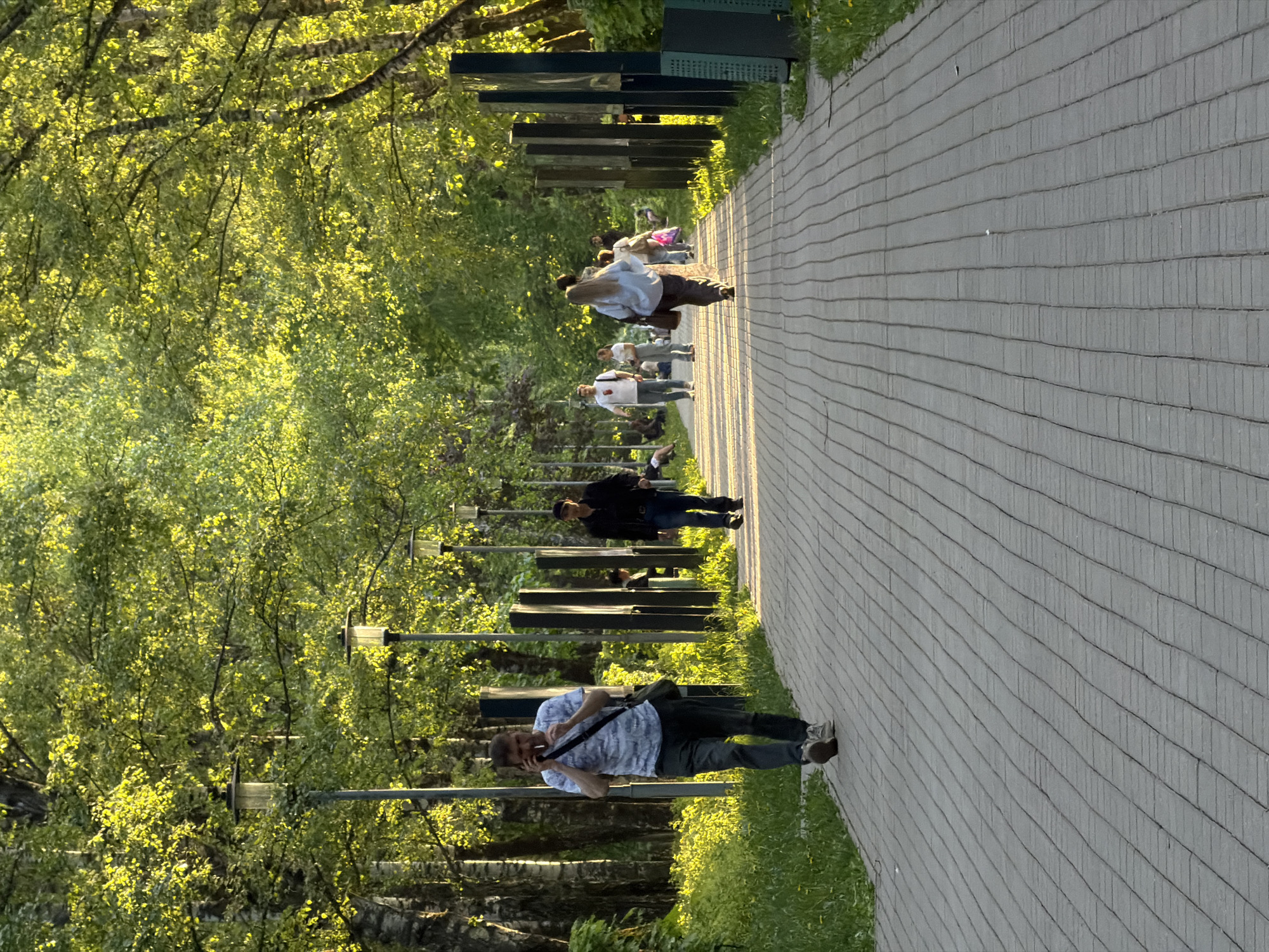 The image captures a park scene in Khimki, Russia. A paved walkway stretches into the distance, lined on one side by trees with vibrant green foliage. Several people are present on the walkway, some walking and others appearing to be resting or observing. Along the path, there are a series of dark, rectangular structures resembling benches or modern art installations. Street lamps are visible interspersed amongst the trees. In the foreground, a man stands holding a phone, possibly taking a picture. The scene is bathed in sunlight, creating strong contrasts and highlighting the textures of the trees and pavement.