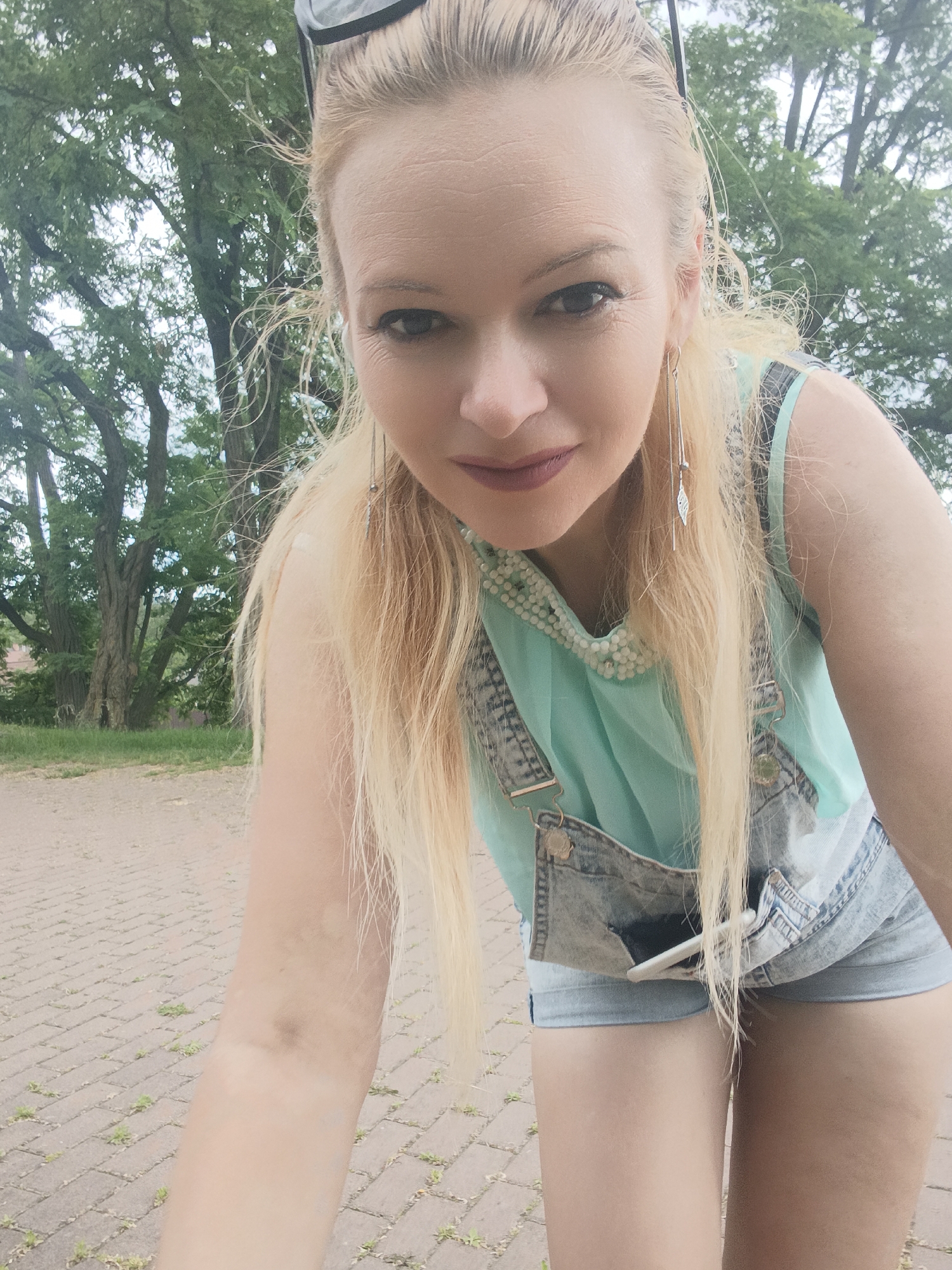 A woman with blonde hair and makeup is taking a selfie outdoors. She is wearing denim overalls over a light blue top and has long, dangly earrings. She is leaning forward, and her face is the primary focus of the image. The background shows trees and a brick pathway. The scene appears to be a park or an outdoor public space in Chernihiv, Ukraine.