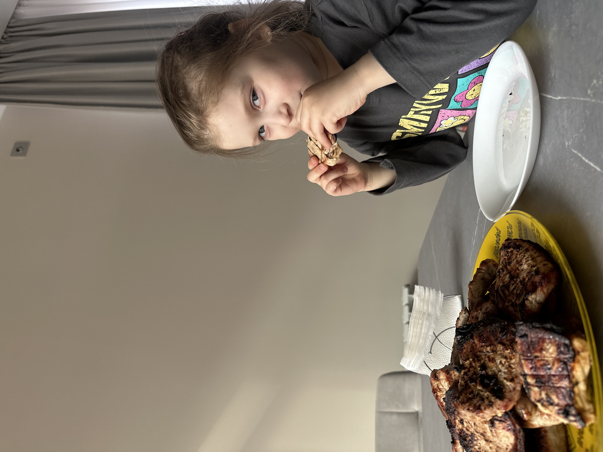 An indoor, vertically oriented shot, taken in Chaikovsky, Russia, features a young girl eating at what appears to be a dining table. The girl, with light brown hair pulled back, is seen from the chest up, looking towards the camera while holding a piece of grilled meat in both hands and bringing it to her mouth. She is wearing a dark long-sleeved shirt with colorful, patterned graphics on the front.
In front of her, on a grey countertop or table, there is a white oval-shaped plate. To her right, a larger yellow plate holds several pieces of dark, grilled meat, possibly pork chops, some with visible grill marks. A white napkin holder with white folded napkins sits between the two plates. Behind the table, light-colored walls and dark grey horizontal blinds or curtains are visible. A small, square light fixture is also mounted on the wall in the upper left background. The text "FOLIA" and "pepperon" (or similar) is partially visible on the rim of the yellow plate. The girl appears focused and content, enjoying her meal.