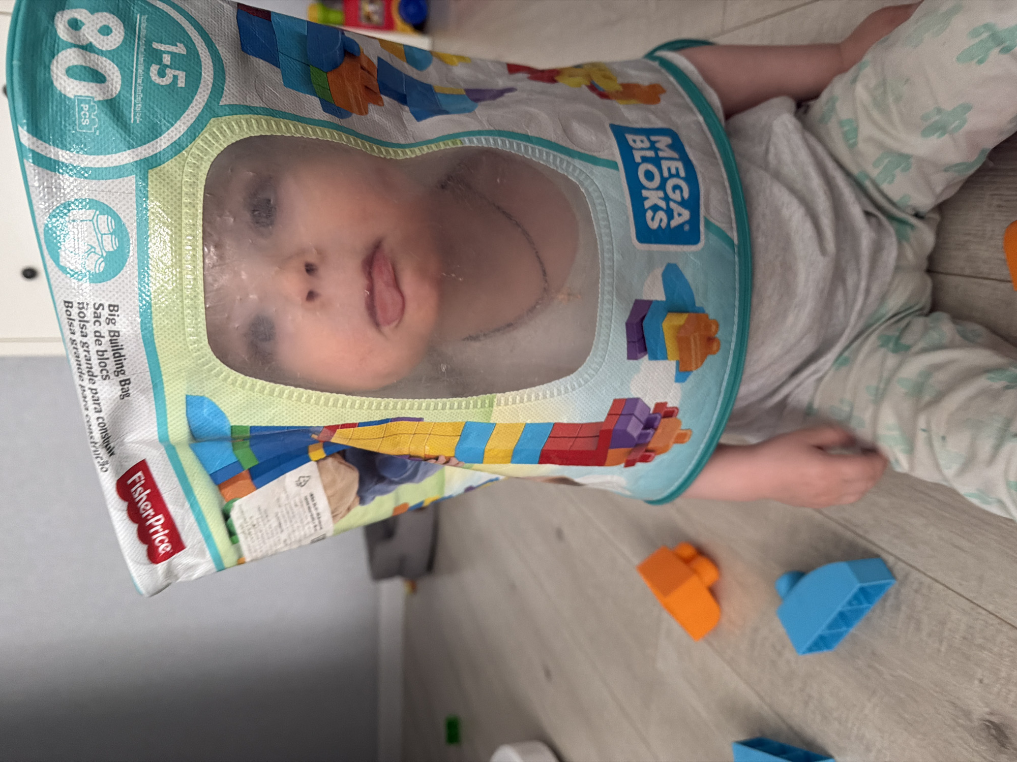 A baby lies on their back on a light-colored, wood-look floor. The baby wears light grey pajamas with a cactus pattern and a light grey t-shirt. A Fisher-Price Mega Bloks bag covers the baby's head. An orange and blue building block are next to the child. The bag features the text "MEGA BLOKS," "1-5," "80 PCS," "Big Building Bag," "Sac de blocs," "Bolsa grande para construir," and "Fisher Price." The baby's tongue is sticking out. The location is not discernible to be in Votkinsk, Russia.