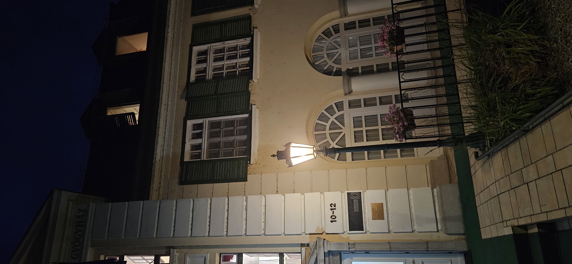 The image shows a nighttime exterior view of a building in Hévíz, Hungary, identified by the signage "GYÓGYHÁZ" (meaning "spa" or "health center") and the address "10-12". The building has a cream-colored facade with white architectural details. The upper stories feature dark-shuttered windows, some of which are illuminated from within. In the foreground, a lit ornate street lamp stands beside a paved walkway. Adjacent to the walkway is a black metal railing with planters holding pink flowers. Beyond the railing, large arched windows with decorative grilles are visible. The ground is a mix of paving stones and a green mat. Lush green foliage and gravel are present near the base of the building and the railing. The sky is dark, indicating nighttime.