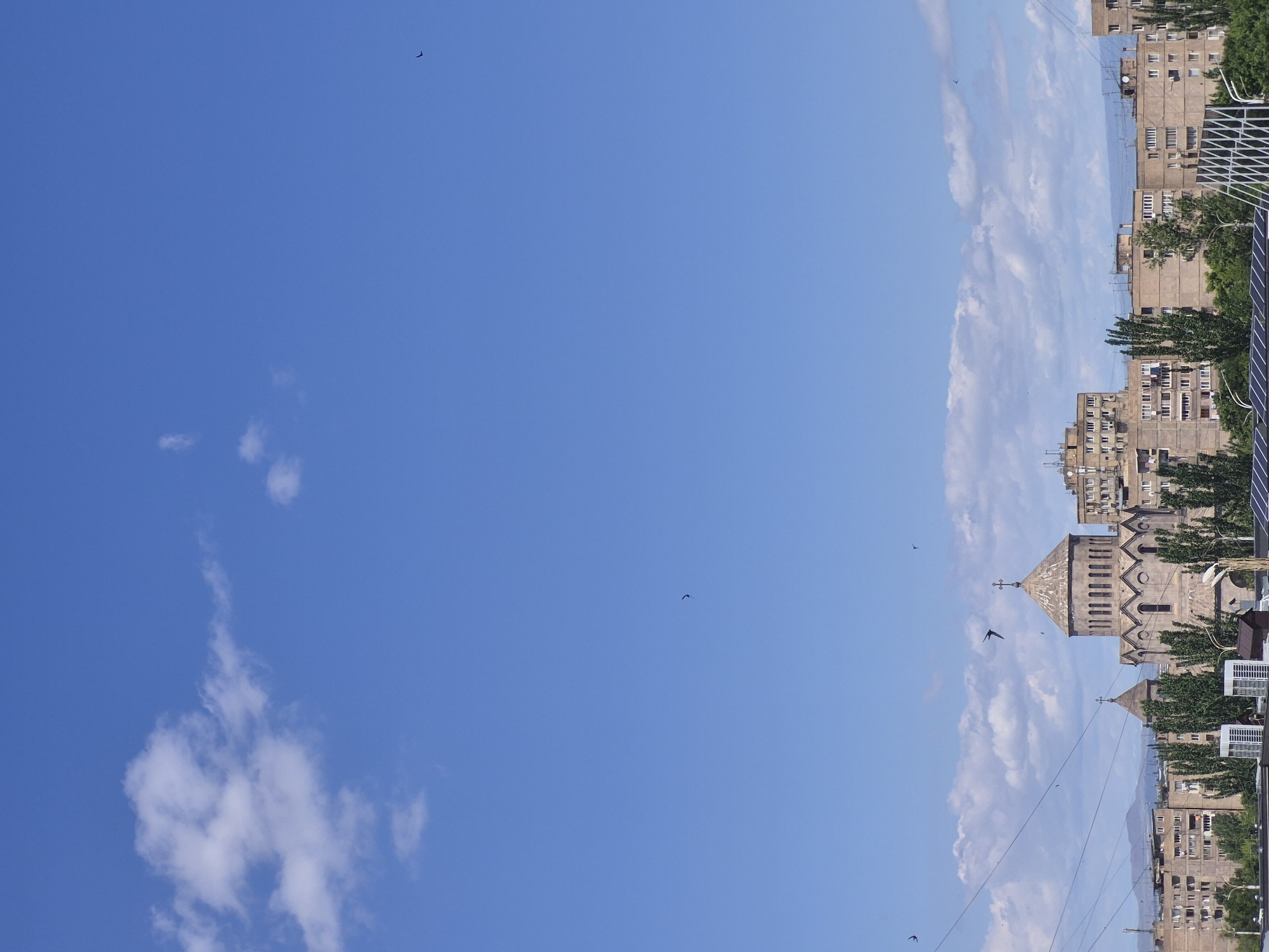 The image depicts a bright, clear daytime scene of an urban environment in Yerevan, Armenia, as indicated by the context. The photograph is oriented vertically but captures a landscape view, with the horizon line running horizontally across the right side.
The majority of the left and central portion of the image is dominated by a vibrant blue sky, mostly clear with scattered, wispy white clouds and some smaller, puffier formations. Several small, dark silhouettes of birds are visible against the blue sky, appearing to be in flight.
On the right side, a cityscape stretches across the horizon. Multiple multi-story buildings, constructed from light-colored stone (characteristic of Yerevan's tuff architecture), are visible. These appear to be residential or apartment blocks with numerous windows. Prominently featured among these buildings is a distinctive structure resembling an Armenian church or monastery, with a tall, polygonal drum topped by a conical dome and adorned with a cross. This architectural style is a strong cultural indicator of the region.
Green trees with lush foliage are interspersed between the buildings, providing a touch of nature within the urban setting. Thin utility lines or power cables are visible stretching across the mid-ground against the sky. On the far right, a partial view of a metal grating or structure, possibly part of a fence, balcony, or solar panel installation, can be seen.
The bright blue sky and clear lighting indicate it is daytime, likely mid-morning or afternoon, with fair weather conditions. There is no discernible human activity or visible text in the image.