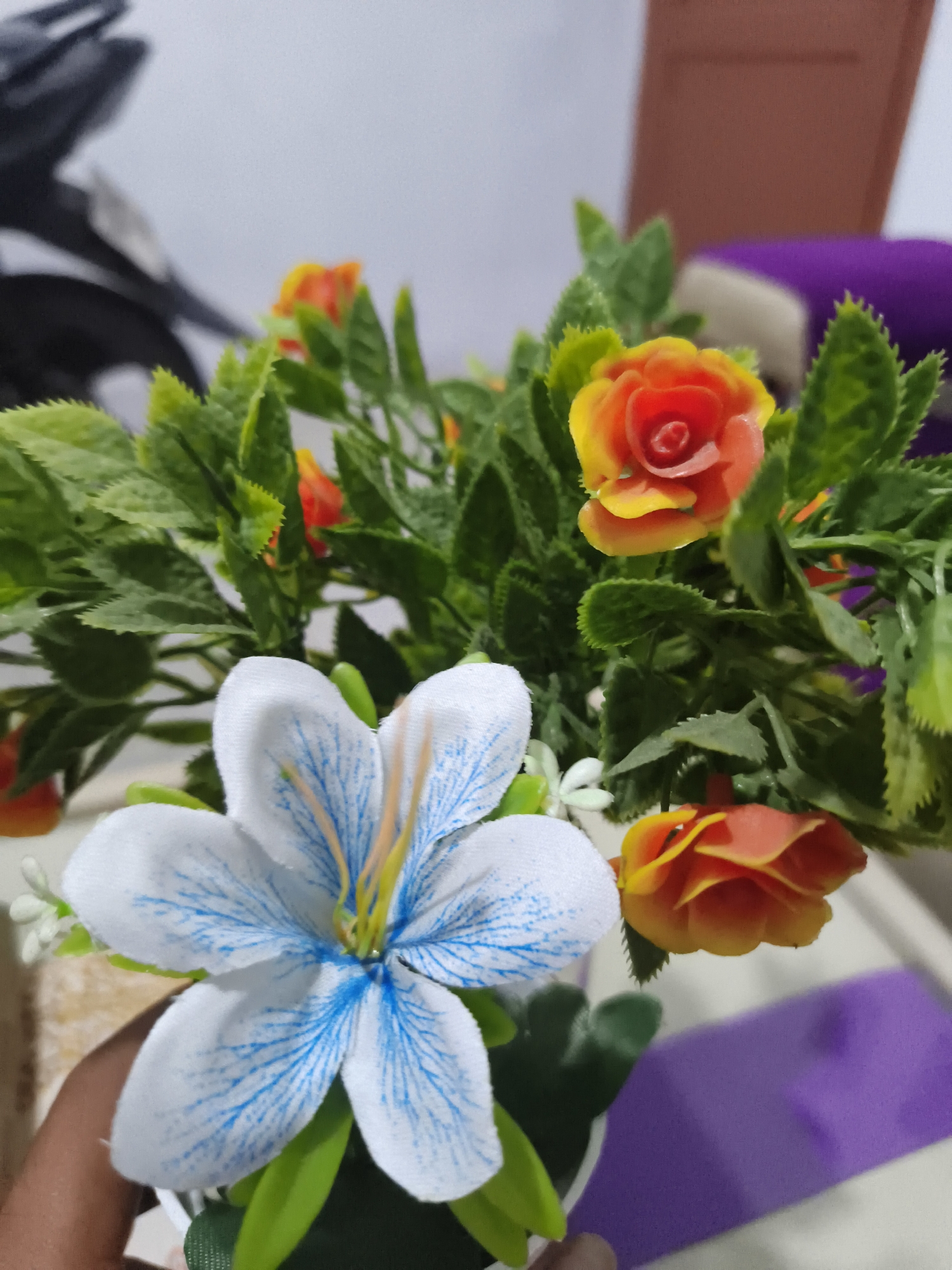 A close-up, eye-level shot captures an arrangement of artificial flowers. In the foreground, a large white flower with blue veins is prominent. Behind it, a cluster of smaller orange and yellow roses with green leaves fills the frame. The background is blurred, showing indistinct objects including what appears to be a motorcycle on the left and a purple object on the right. The scene is likely indoors, given the lighting and the presence of indoor objects. The image does not provide any specific indicators of its location in Pangkalpinang, Indonesia.