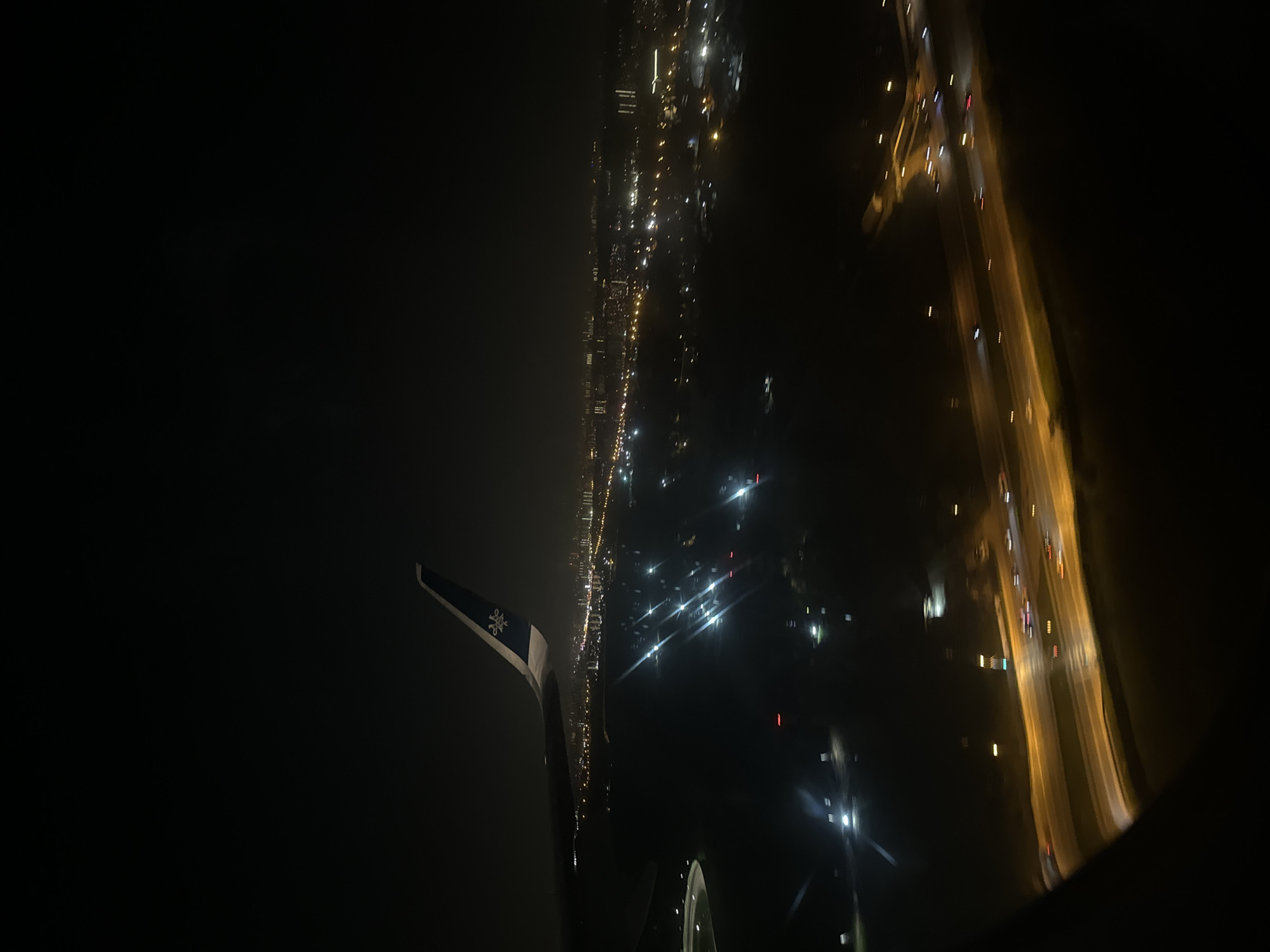 A nighttime scene, likely taken from an airplane window, depicts a cityscape and highway illuminated by artificial lights. On the left, a portion of the airplane wing with a visible emblem is present. The city lights stretch across the middle of the frame, displaying various illuminated buildings. To the right, a blurred highway with the streaks of moving vehicles is visible. It depicts Khimki, Russia at night.