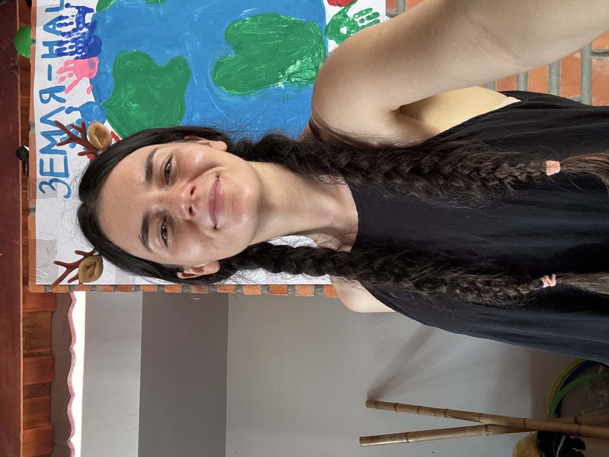 A woman with long dark hair in two braids stands in front of a painted poster, likely in the city of Mũi Né, Vietnam. The poster, leaning against a brick structure, depicts a green and blue Earth with what appears to be handprint art and the word "ЗЕМЛЯ-НАШ" written vertically on the left. The woman is wearing a black top and has a small decorative item in her hair. Two bamboo poles rest near the base of a grey and white wall in the lower part of the frame. The scene appears to be indoors.