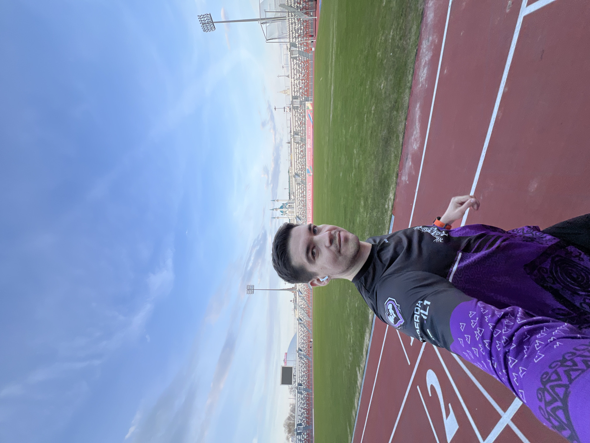 A medium-shot, eye-level portrait shows a man standing on a red track next to a green field with stadium seating. The man has short brown hair and is wearing a black and purple long-sleeved shirt. He is looking directly at the camera with a slight smile, wearing a white wireless earbud in his left ear. In the background is a stadium with seating and floodlights. Beyond the stadium, the cityscape of Kazan, Russia is visible. The sky above is blue with light clouds.