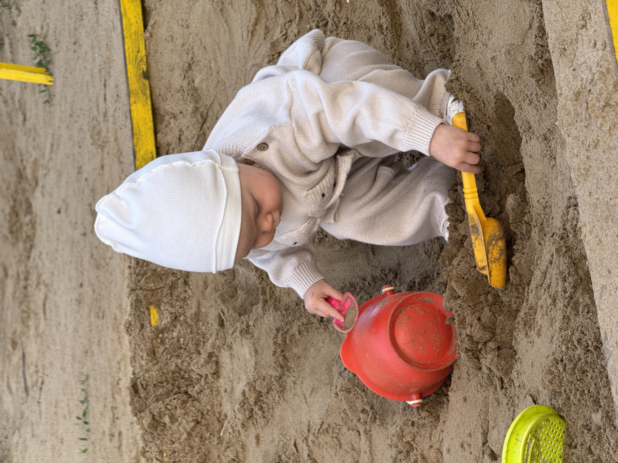 A baby wearing a white hat and a cream-colored outfit is playing in a sandbox filled with sand. The baby is focused on a red toy teapot and a yellow toy shovel. The sandbox has yellow borders, one of which is visible on the left side of the image. There is also a bright green toy in the lower right corner. The sand appears dry and is piled up in various mounds. The lighting suggests it is daytime, possibly outdoors given the sand and toy context. The baby appears engrossed in their play. No text is visible in the image. The location is identified as Vladimir, Russia.
